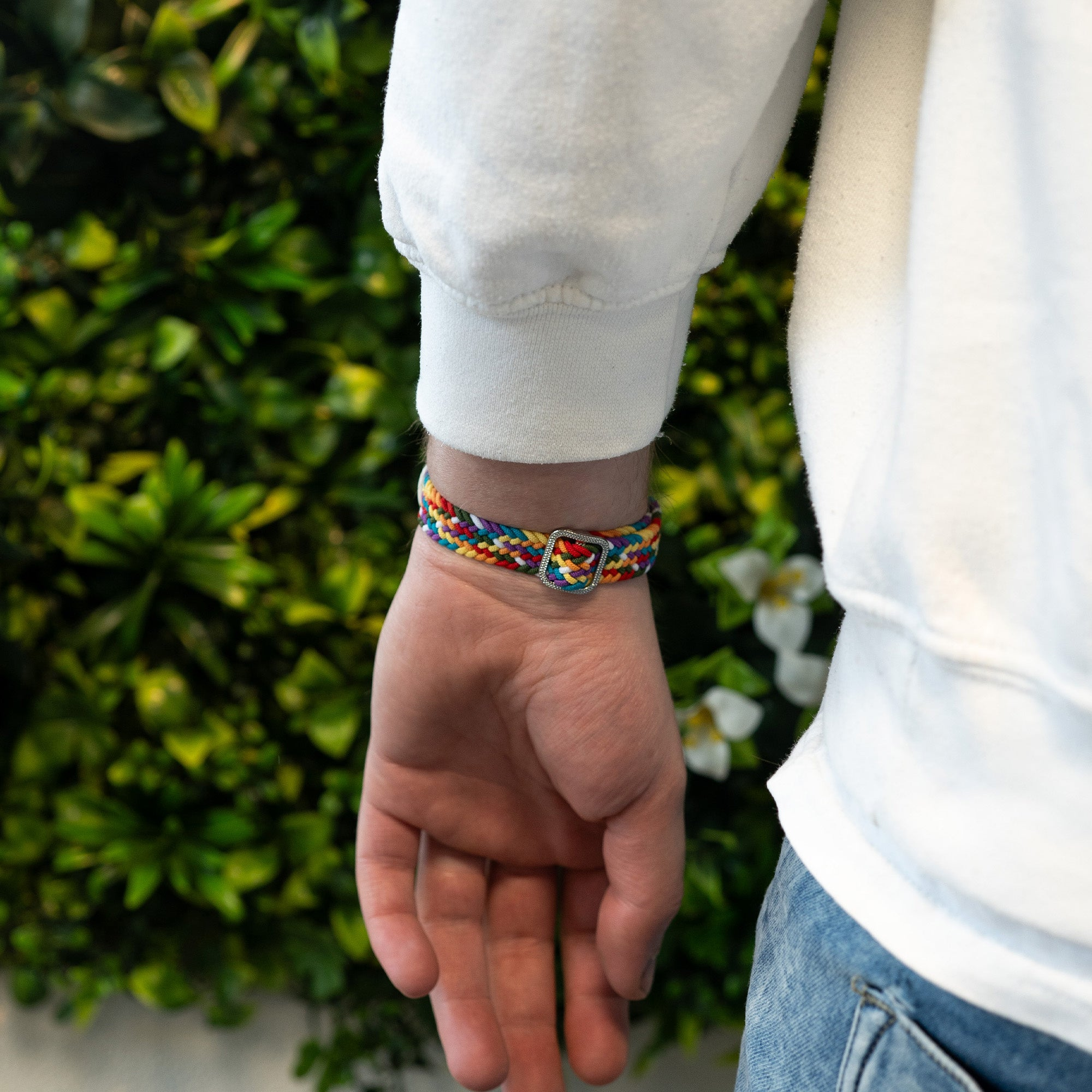 Das Handgelenk einer Person mit einem farbenfrohen geflochtenen Armband. Ein weißer Pullover und grünes Laub im Hintergrund.