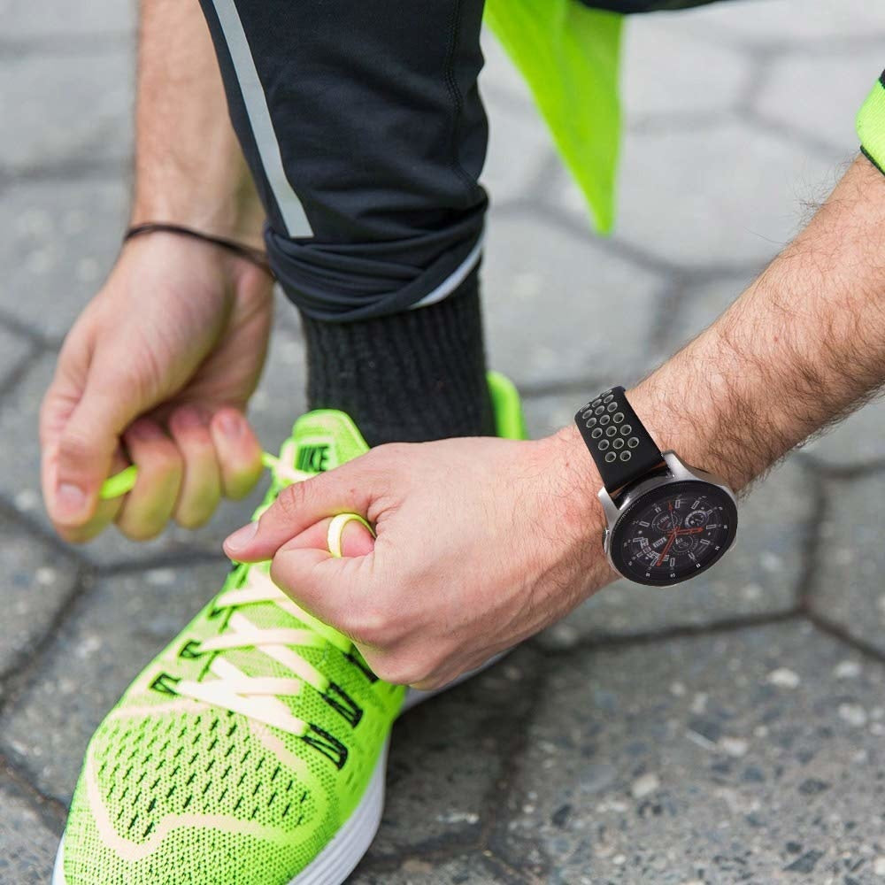 Eine Person bindet einen neongrünen Laufschuh. Sie trägt eine Uhr mit schwarzem Armband. Dunkle Hose und hellgrünes Hemd sind sichtbar.