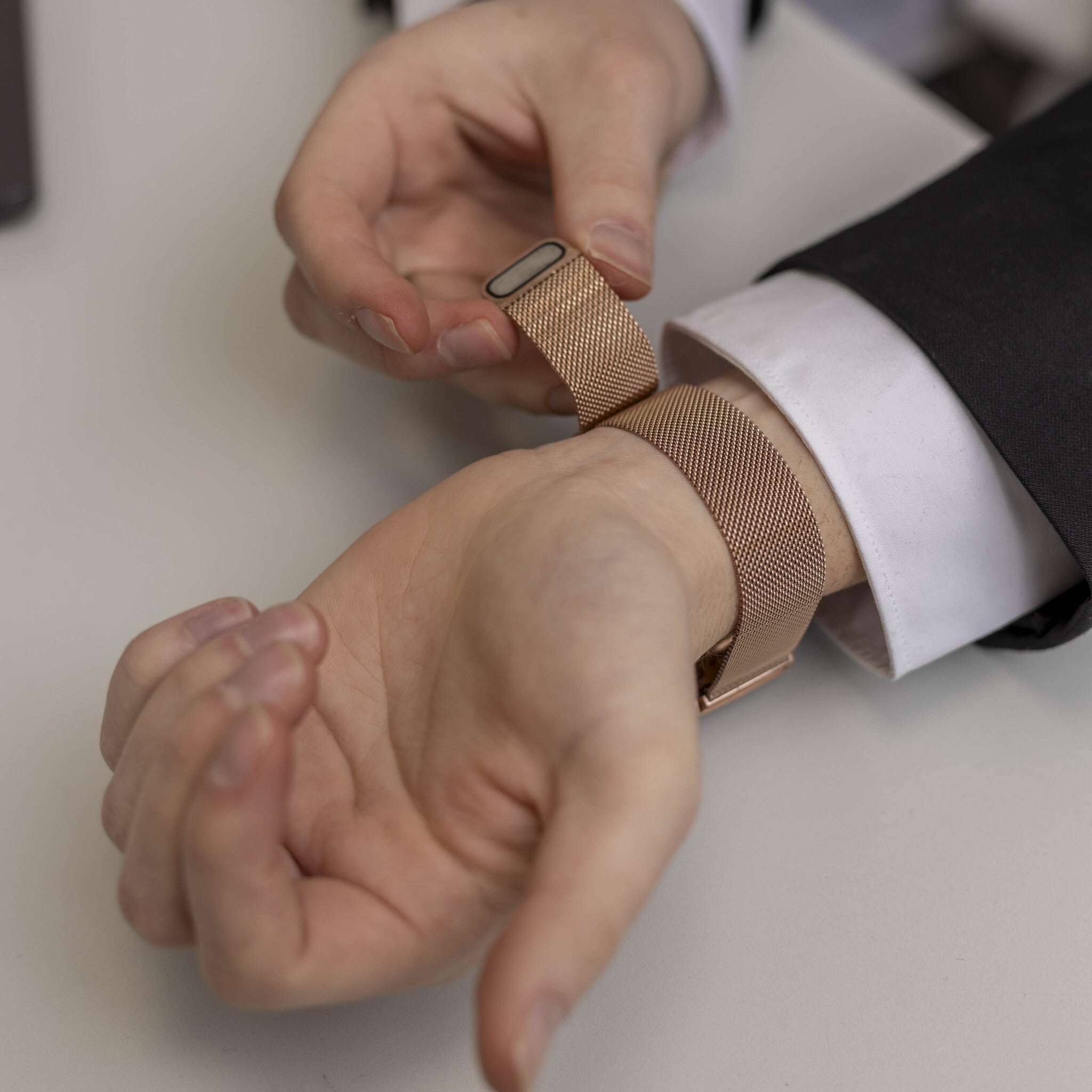 Eine Person befestigt eine goldene Uhr an ihrem Handgelenk. Die Uhr hat ein Mesh-Armband. Sie tragen einen Anzug.