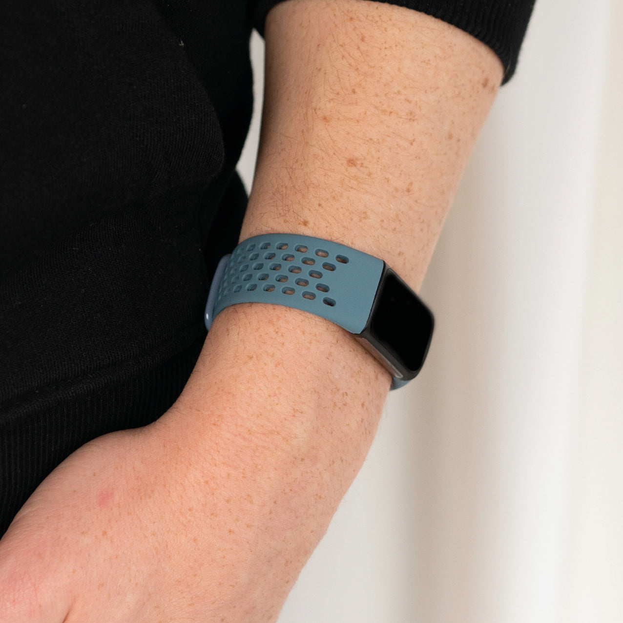 Arm einer Person mit Sommersprossen, die einen blauen Fitness-Tracker mit schwarzem Bildschirm trägt. Schwarzer Pullover im Hintergrund, weiße Wand.