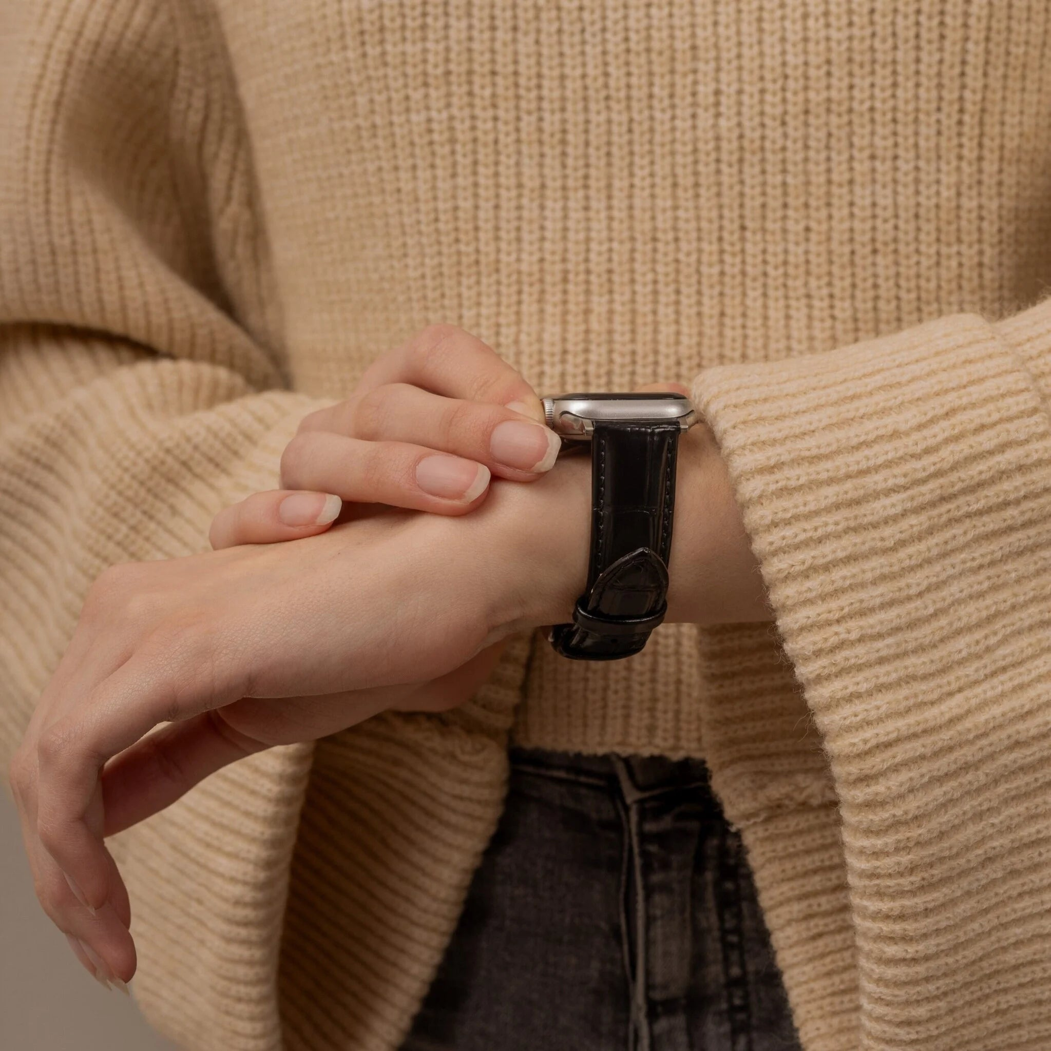 Person trägt einen beigen Pullover und eine schwarze Lederuhr. Hände sind sichtbar. Hintergrund ist ein strukturierter beiger Pullover.