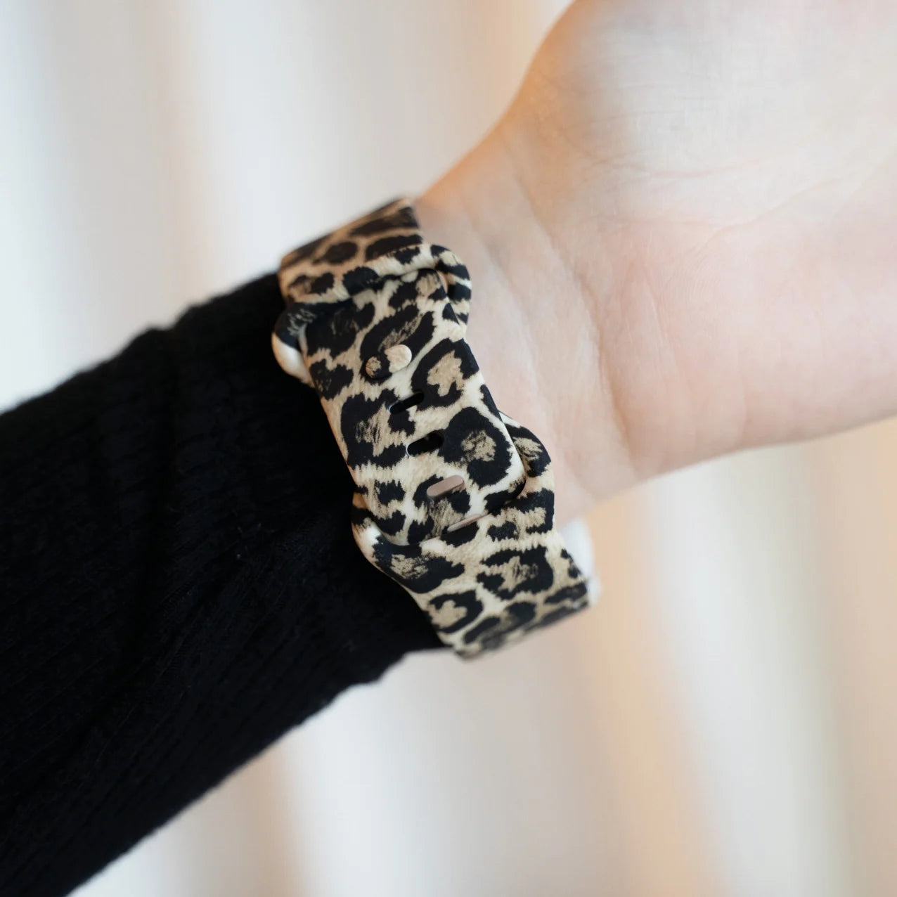 Ein Handgelenk mit einem Armband mit Leopardenmuster. Der Arm trägt einen schwarzen Pullover. Der Hintergrund ist verschwommen und hell.