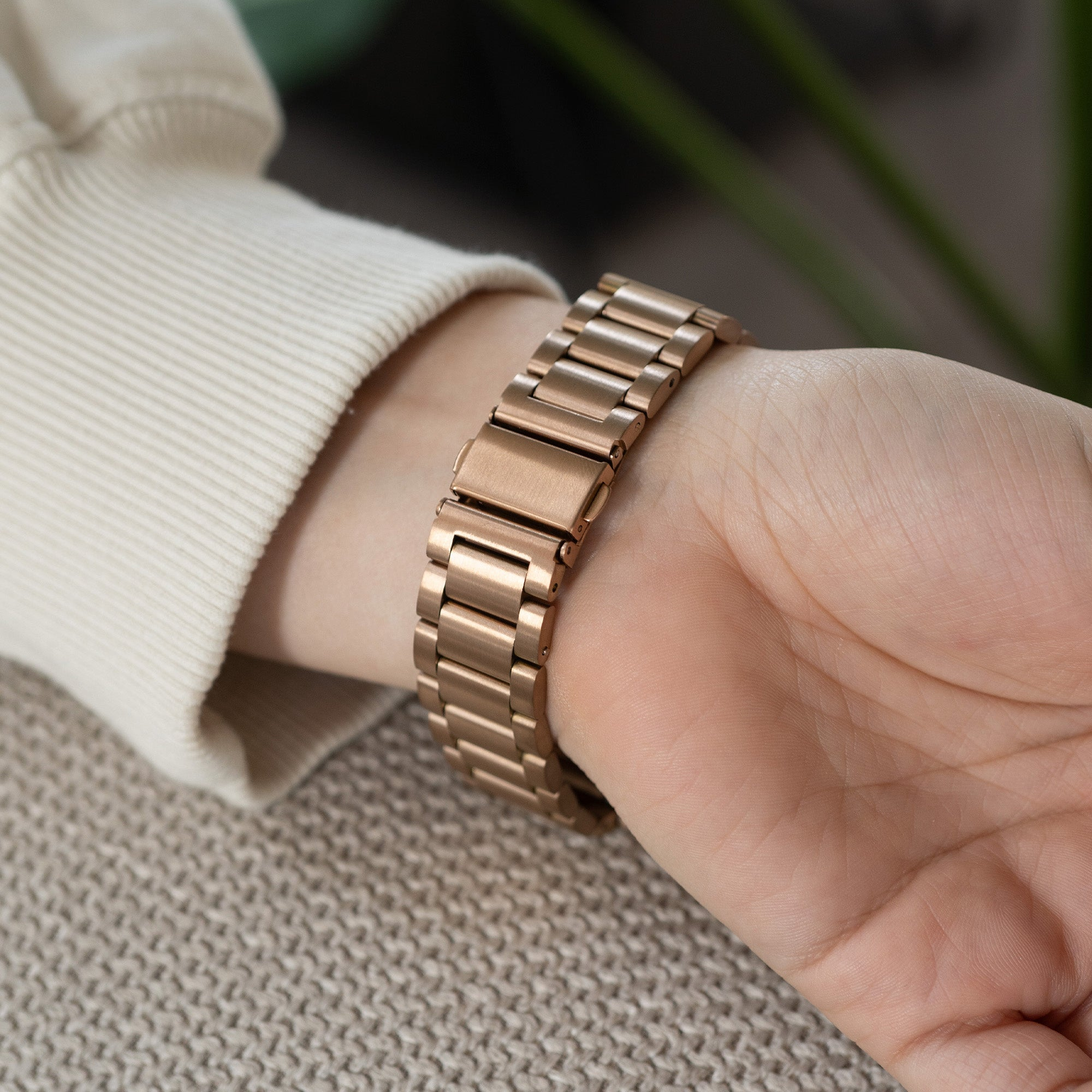 Nahaufnahme eines Handgelenks mit einer roségoldenen Uhr. Auf der linken Seite ist ein cremefarbener Pullover zu sehen.