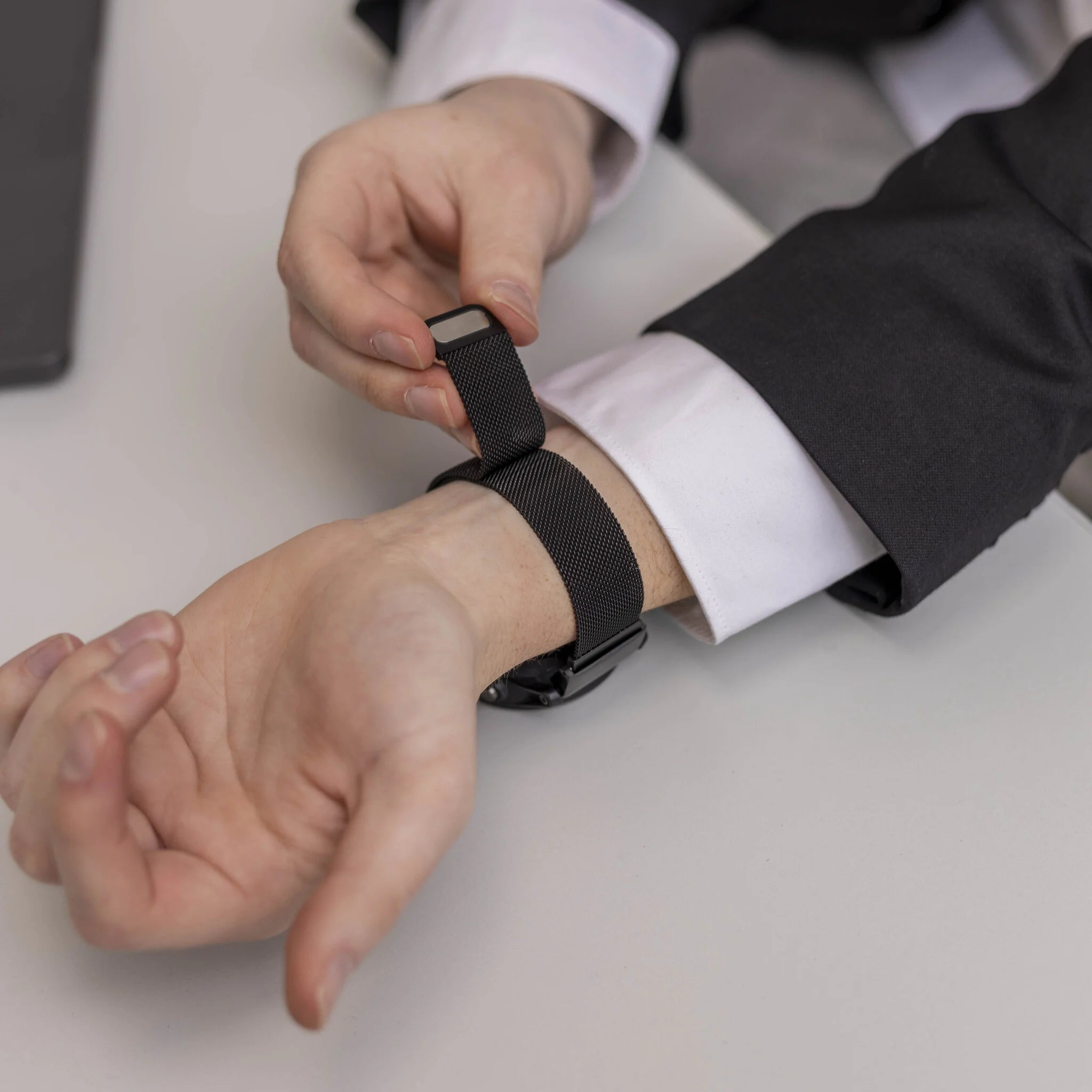 Eine Person im schwarzen Anzug justiert eine schwarze Uhr an ihrem Handgelenk. Die Uhr hat ein Mesh-Armband.