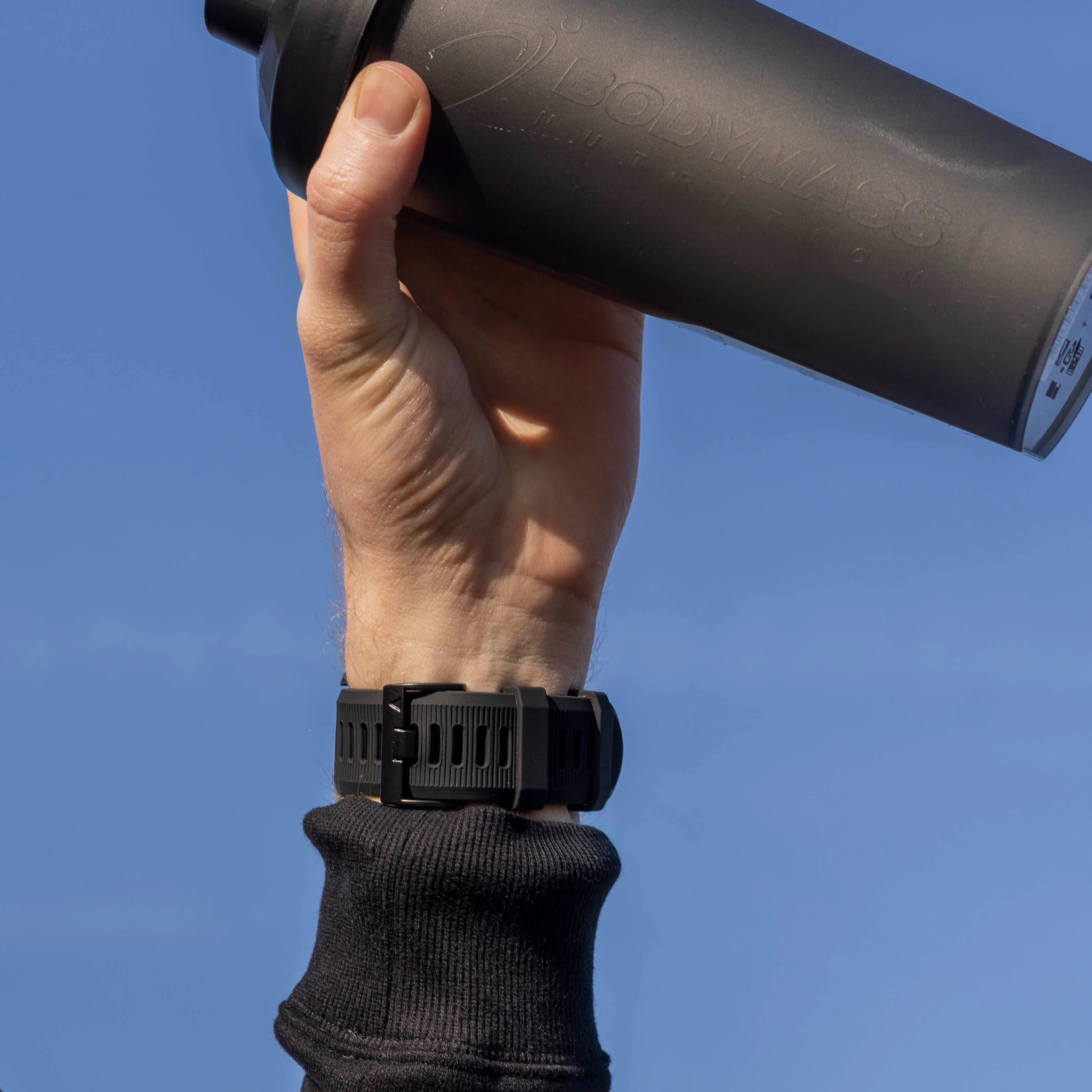 Eine Person hält eine schwarze Shaker-Flasche vor blauem Himmel. Eine schwarze Uhr ist am Handgelenk.