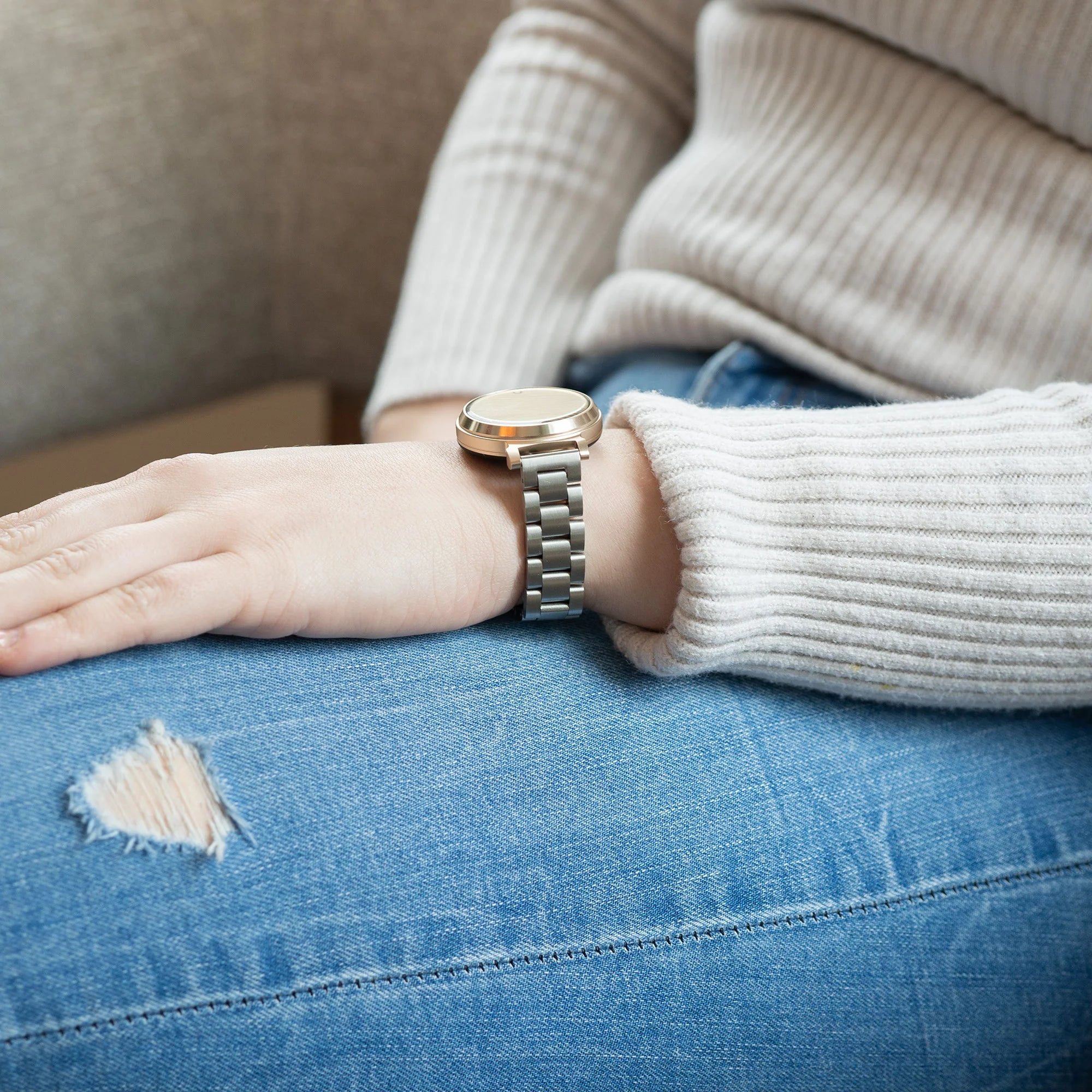 Arm einer Person mit Uhr am Handgelenk. Die Person trägt einen hellen Pullover und zerrissene blaue Jeans.