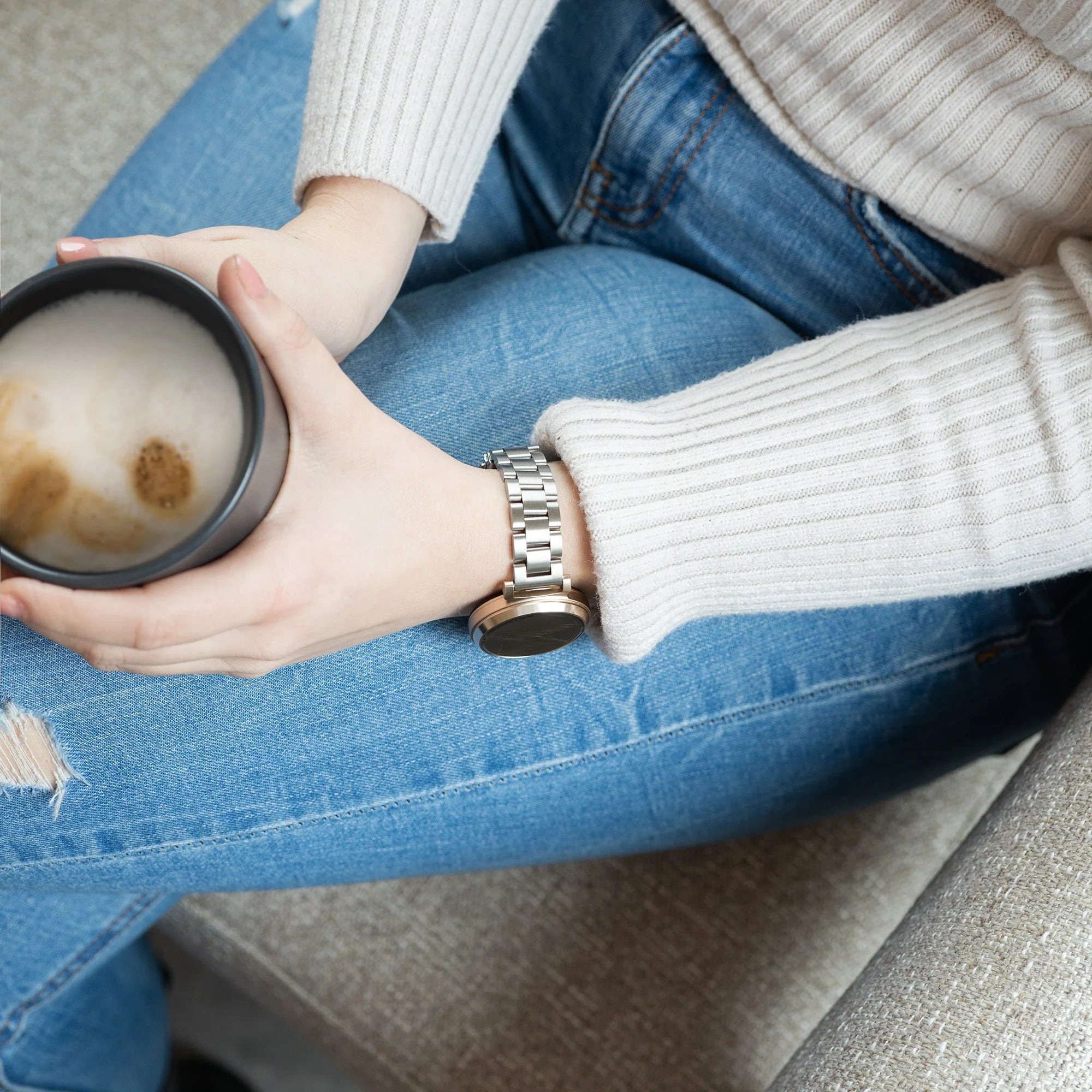 Eine Person hält einen Kaffeebecher. Sie trägt eine Uhr und Jeans. Ein Pullover bedeckt ihre Arme. Der Hintergrund zeigt eine Couch.