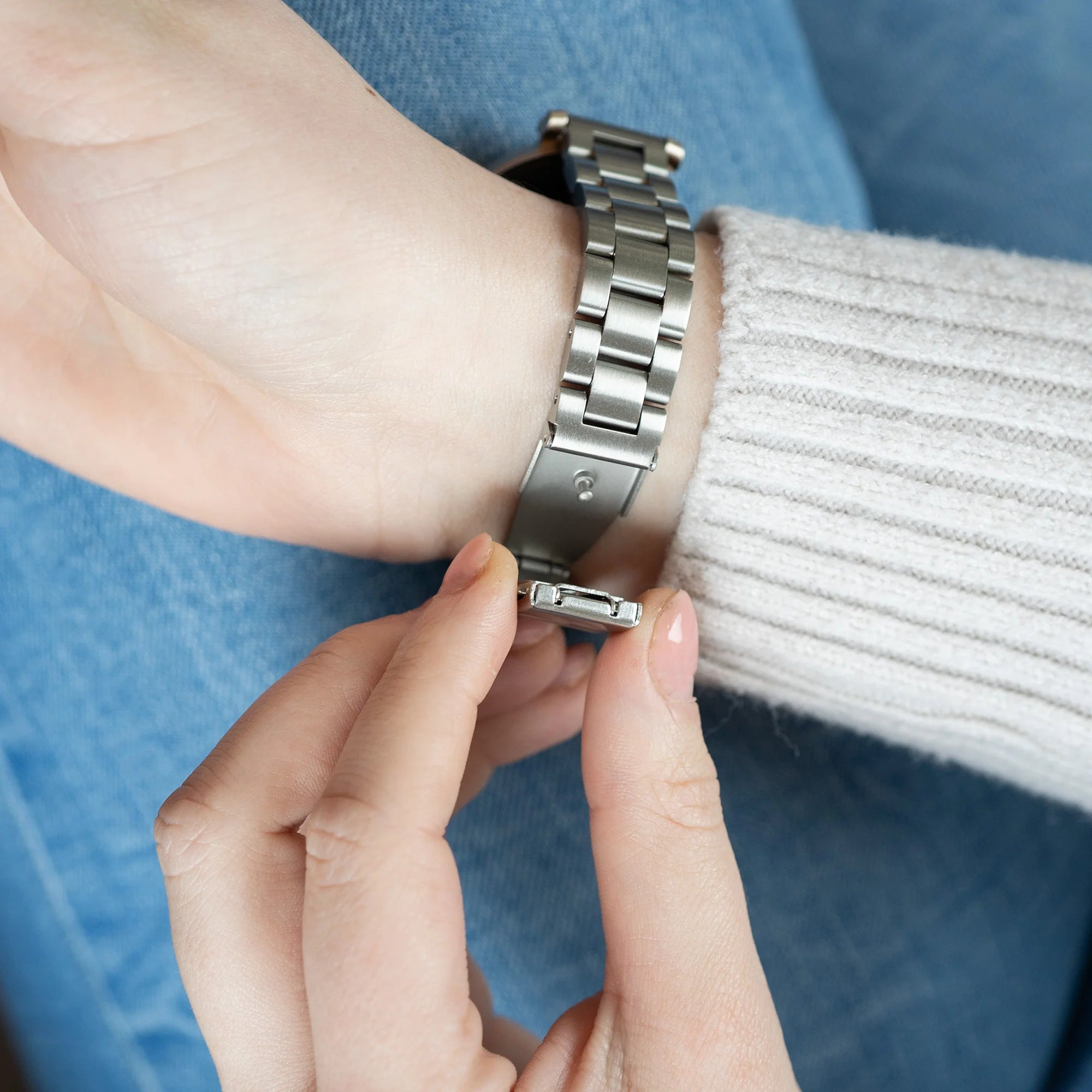 Die Hand einer Person stellt eine silberne Uhr ein. Sie trägt einen grauen Pullover und Jeans. Der Verschluss ist offen.