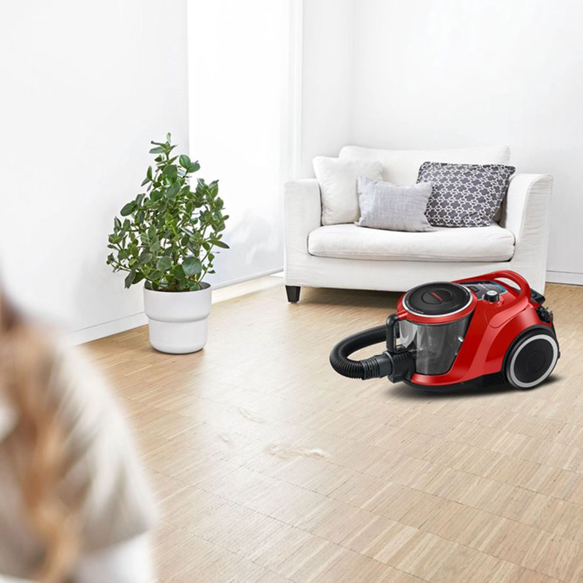 Czerwony odkurzacz na drewnianej podłodze. Biała sofa z poduszkami i doniczkowa roślina w tle.