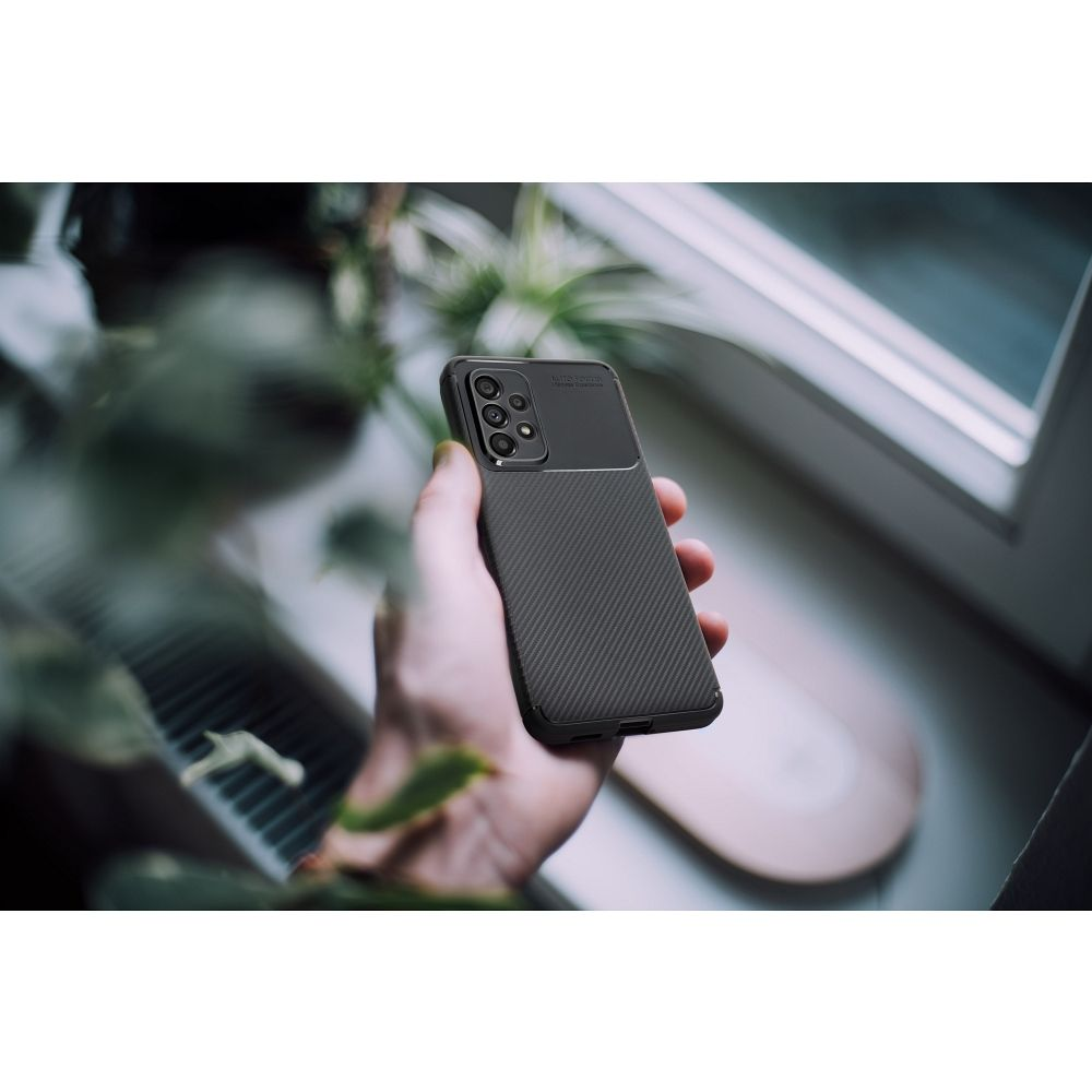 Una mano che tiene un telefono nero con una custodia strutturata. Il telefono ha più fotocamere. Lo sfondo include piante e una finestra.