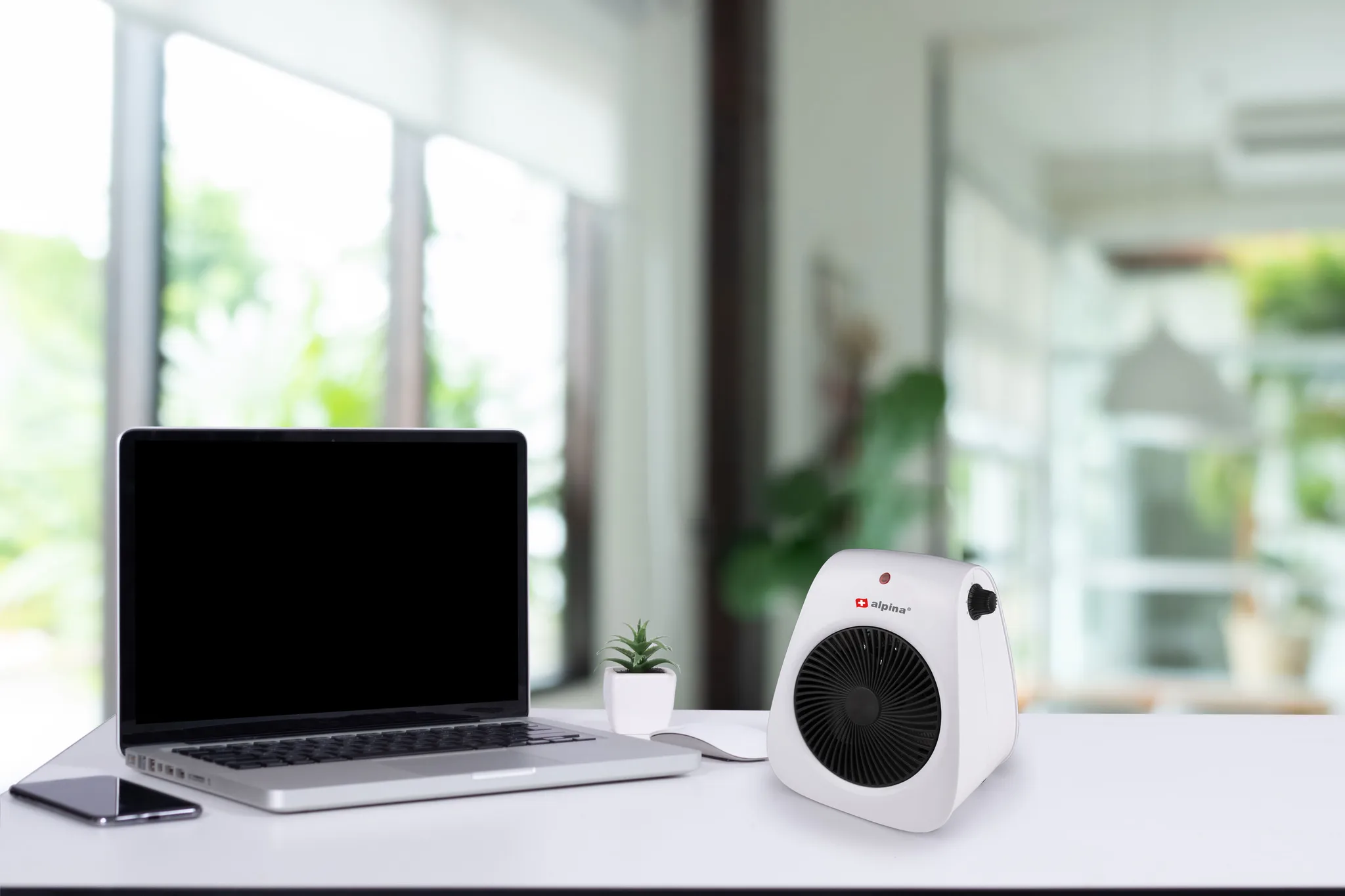 A laptop, a phone, a white heater, and a small plant are on a white desk in front of a window. Background is blurred.