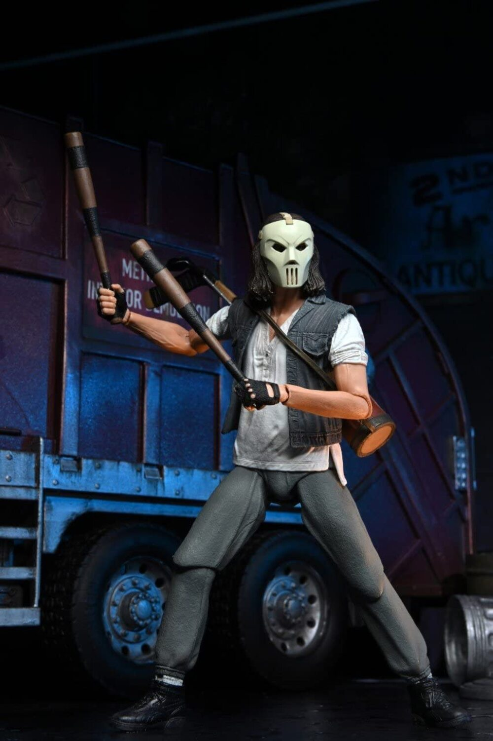 Hokey maskeli ve iki sopalı Casey Jones aksiyon figürü, bir kamyonun önünde duruyor. Yelek ve gri pantolon giyiyor.