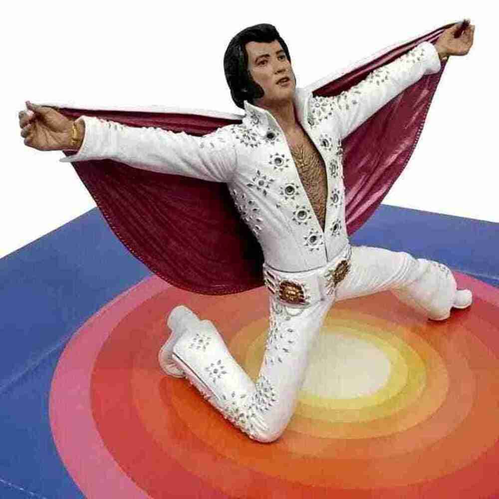 Beyaz tulum ve kırmızı pelerinli bir Elvis heykeli. Renkli bir sahnede diz çökmüş, kolları açık.