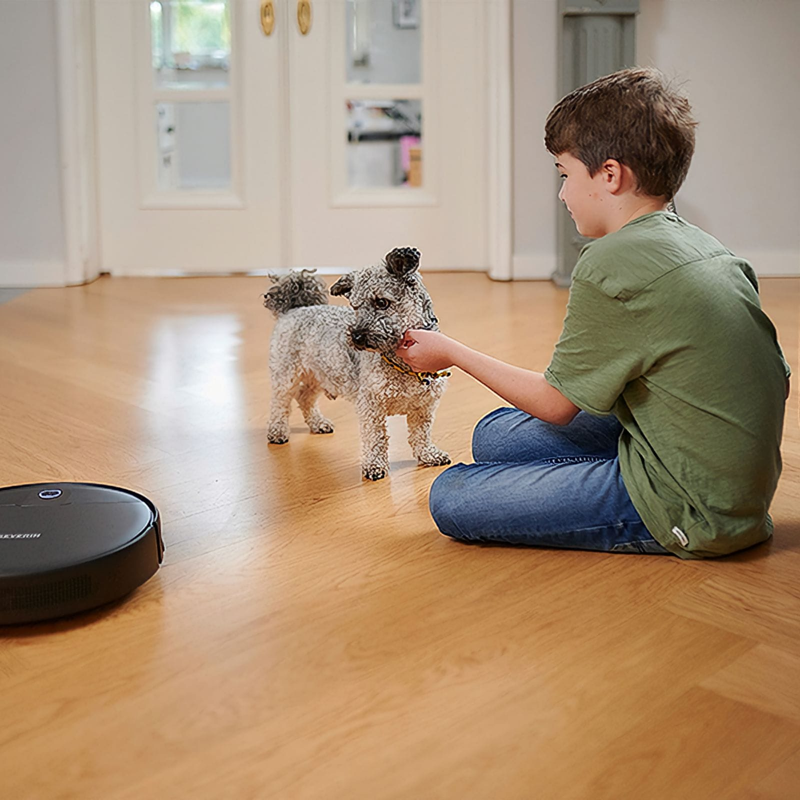 Un ragazzo è seduto per terra che accarezza un cane. Un aspirapolvere robot nero è a sinistra. Pavimento in legno.