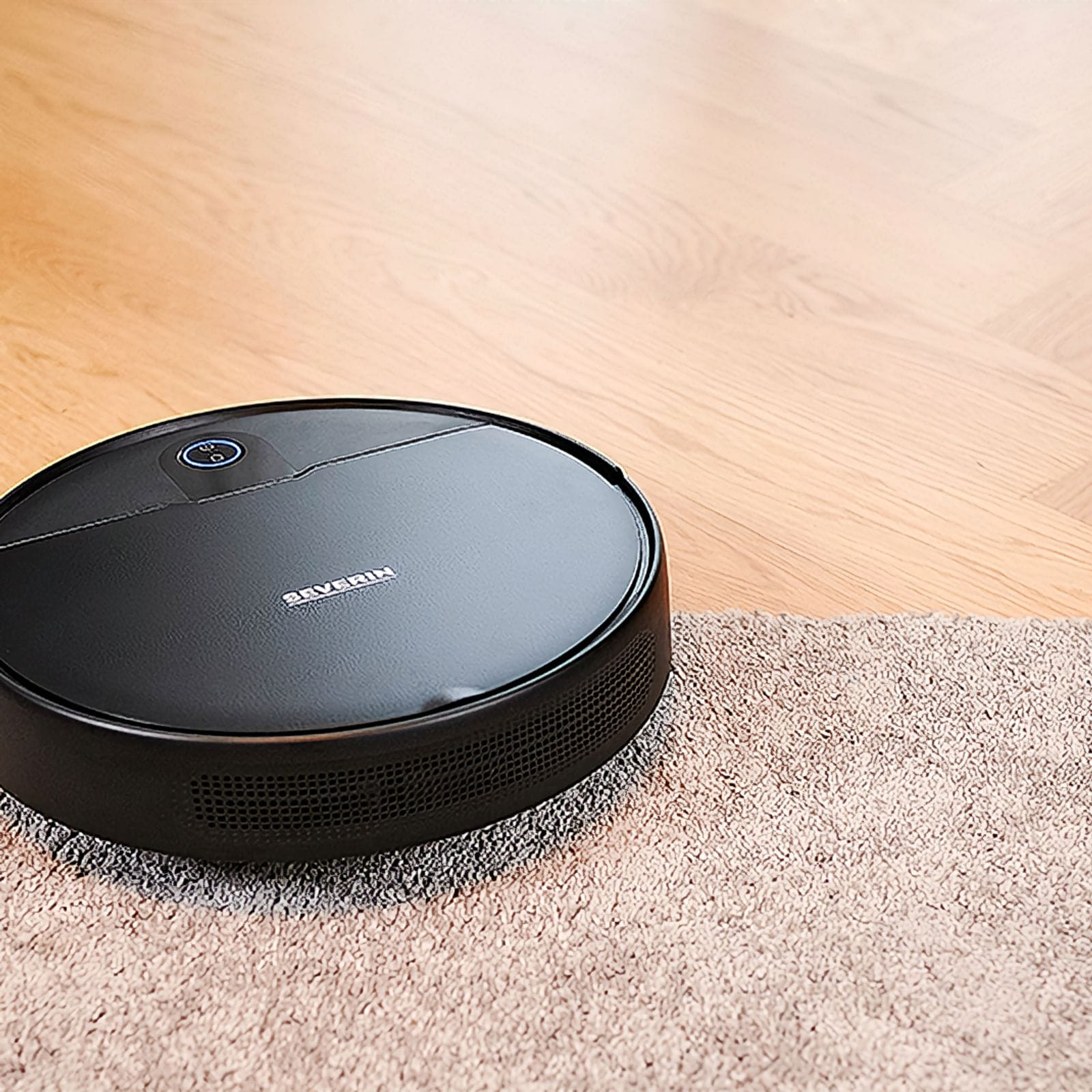 Aspirapolvere robot nero su pavimento in legno, che passa a un tappeto beige. L'aspirapolvere ha il marchio Severin.