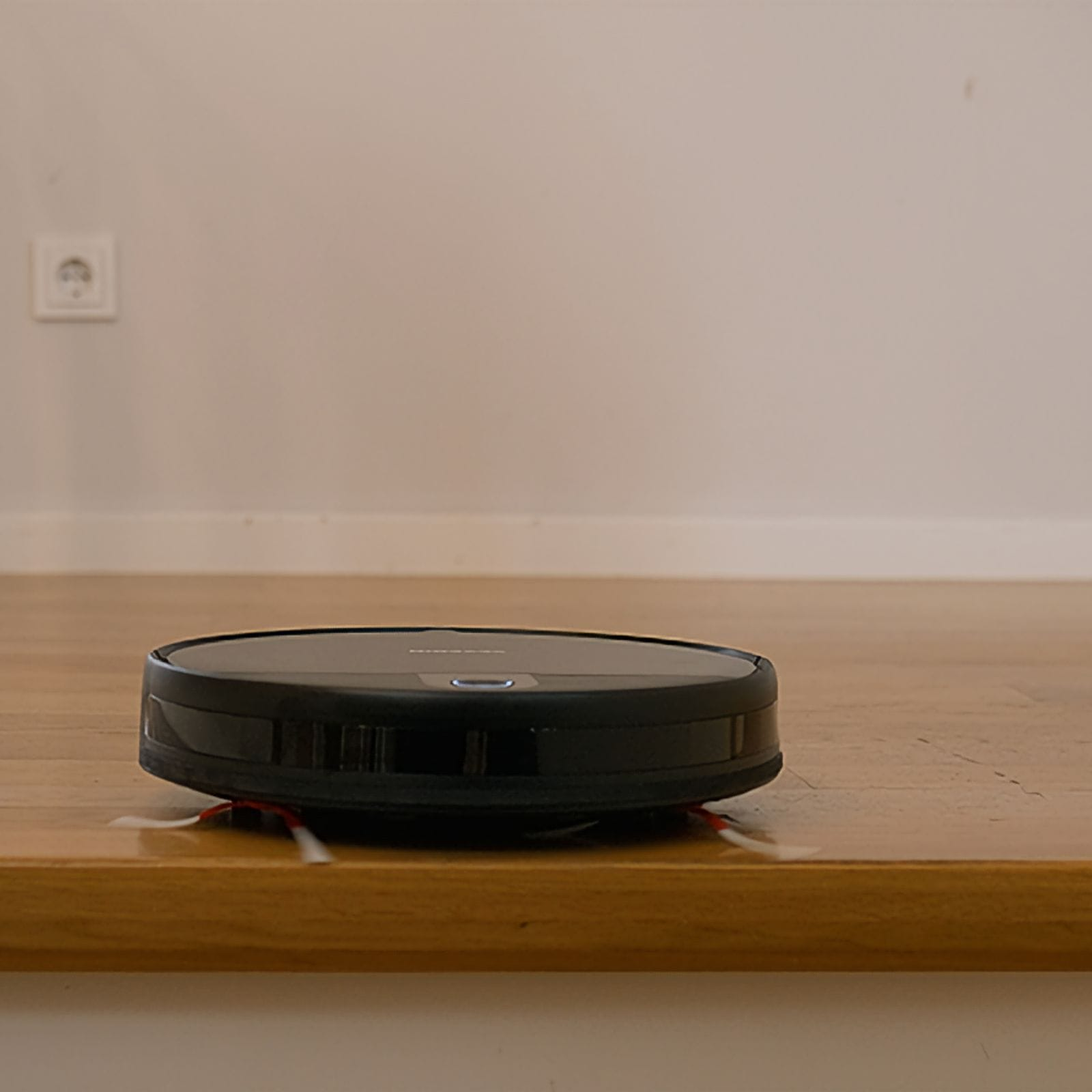 Un aspirapolvere robotico nero su un pavimento in legno. Sullo sfondo un muro bianco e una presa elettrica.