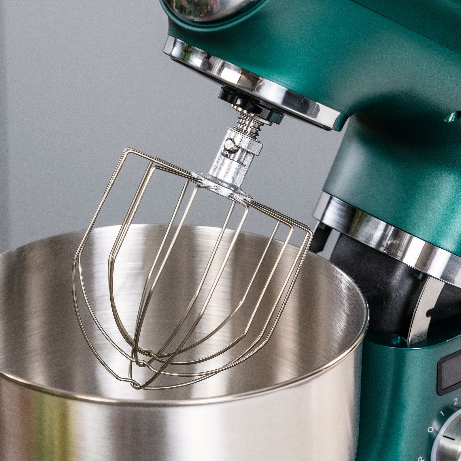 Ein grüner Küchenmixer mit Schneebesenaufsatz befindet sich über einer Edelstahlschüssel. Der Hintergrund ist grau.