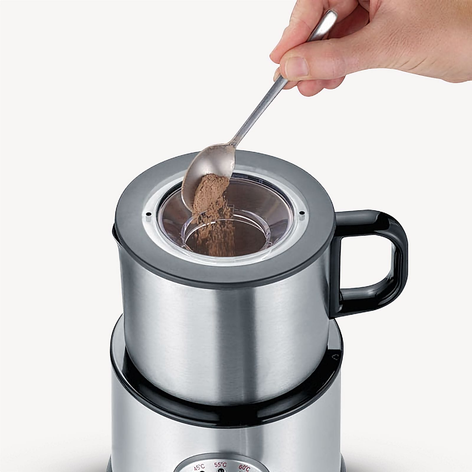 A hand uses a spoon to pour brown powder into a silver machine. Machine has a black handle and base.