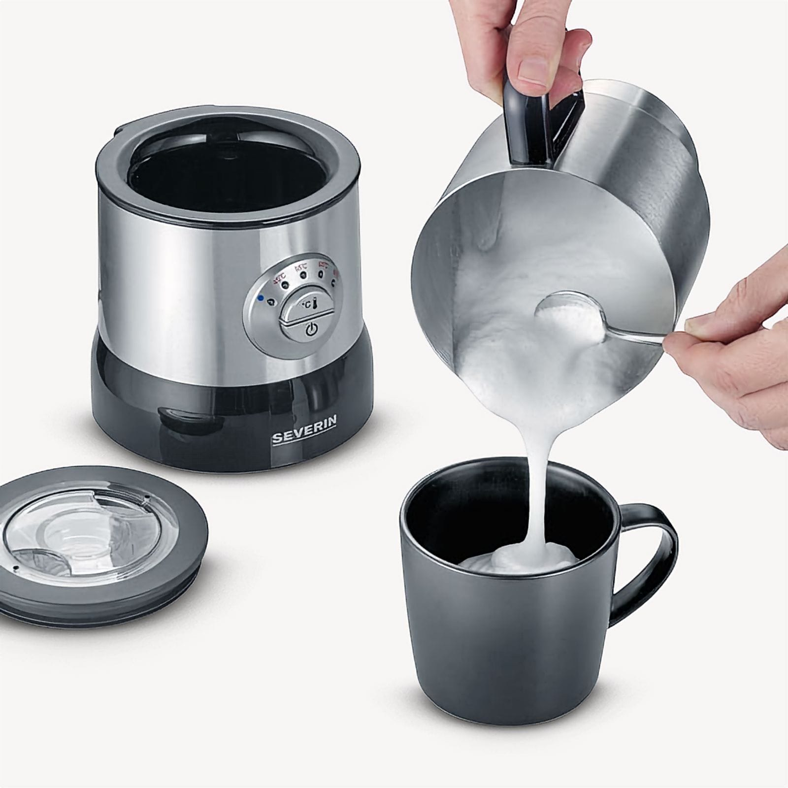 A person pours foamed milk from a stainless steel pitcher into a black mug. A milk frother and its lid are nearby.