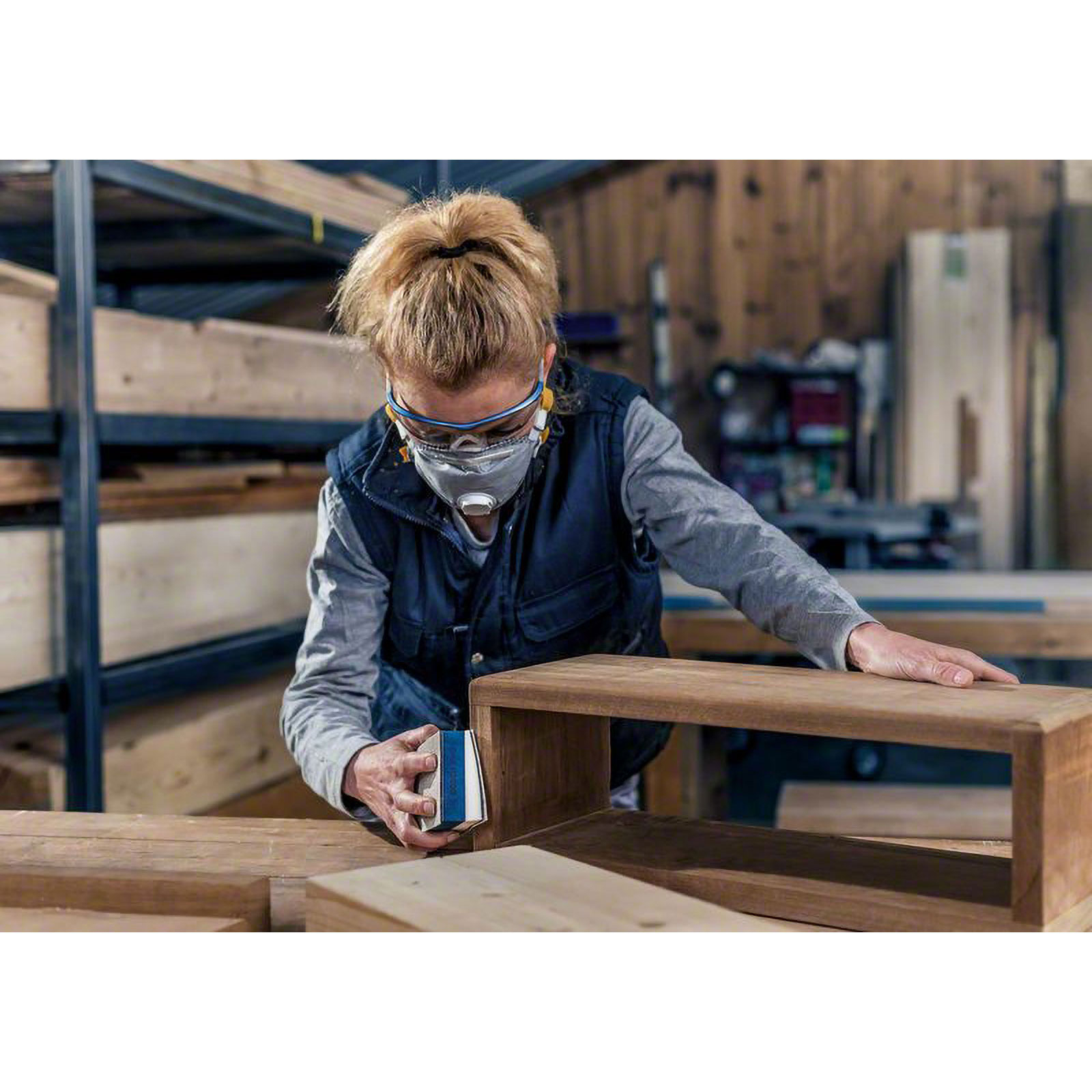 Kobieta szlifuje drewnianą skrzynkę w warsztacie, nosząc maskę i okulary ochronne. W tle półki i drewno.