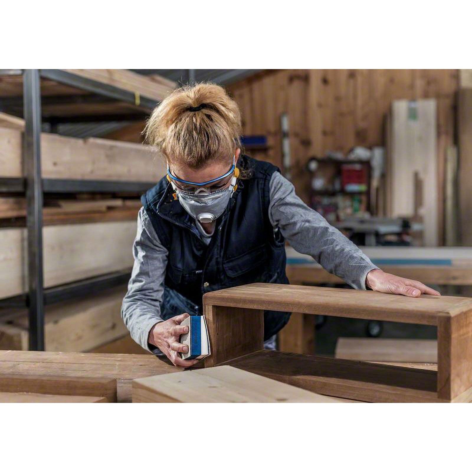 Osoba szlifuje drewnianą skrzynię w warsztacie. Ma na sobie maskę i okulary ochronne. Drewniane półki w tle.