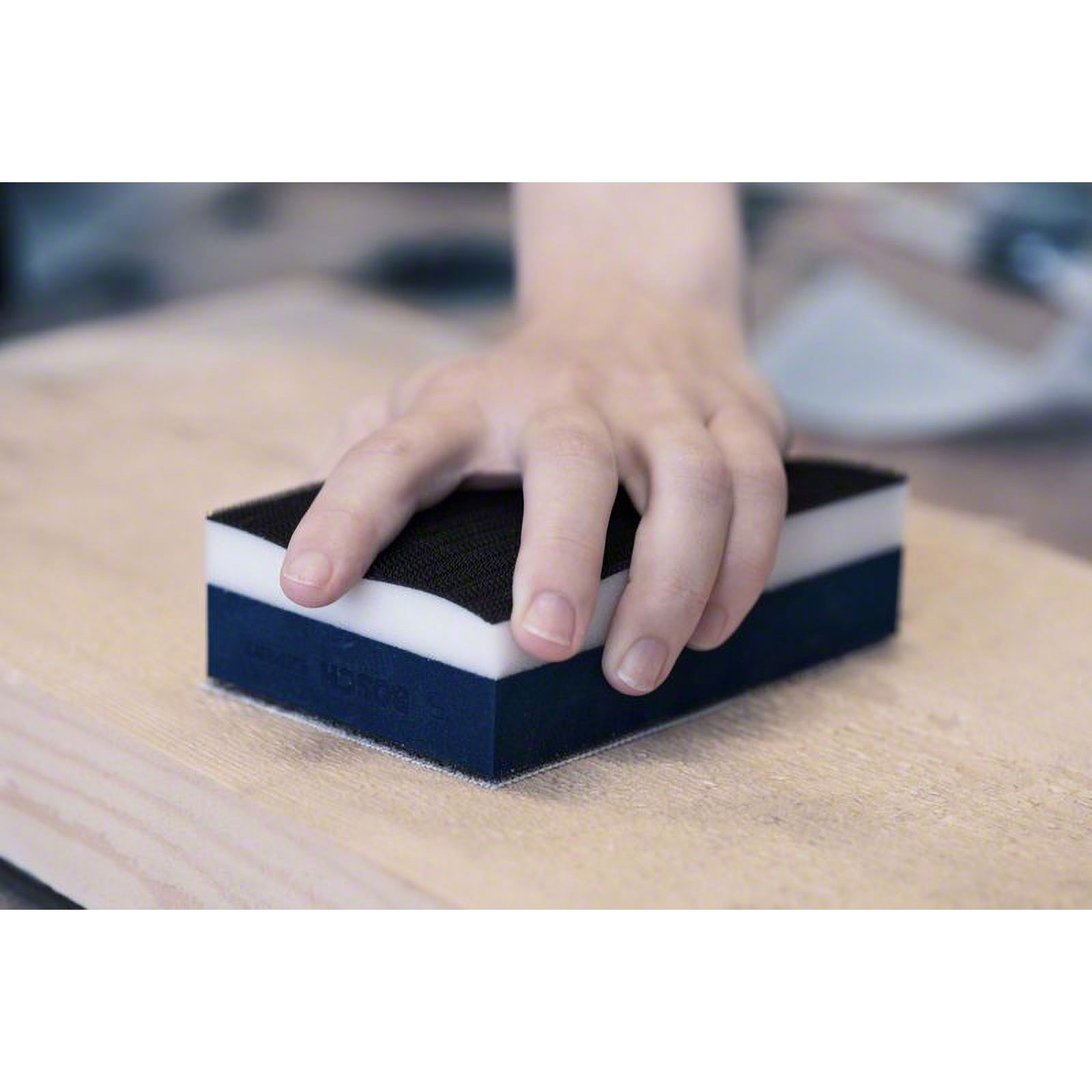 Dłoń trzyma wielowarstwowy klocek szlifierski. Klocek jest czarny, biały i niebieski. Umieszczony na jasnej drewnianej powierzchni.