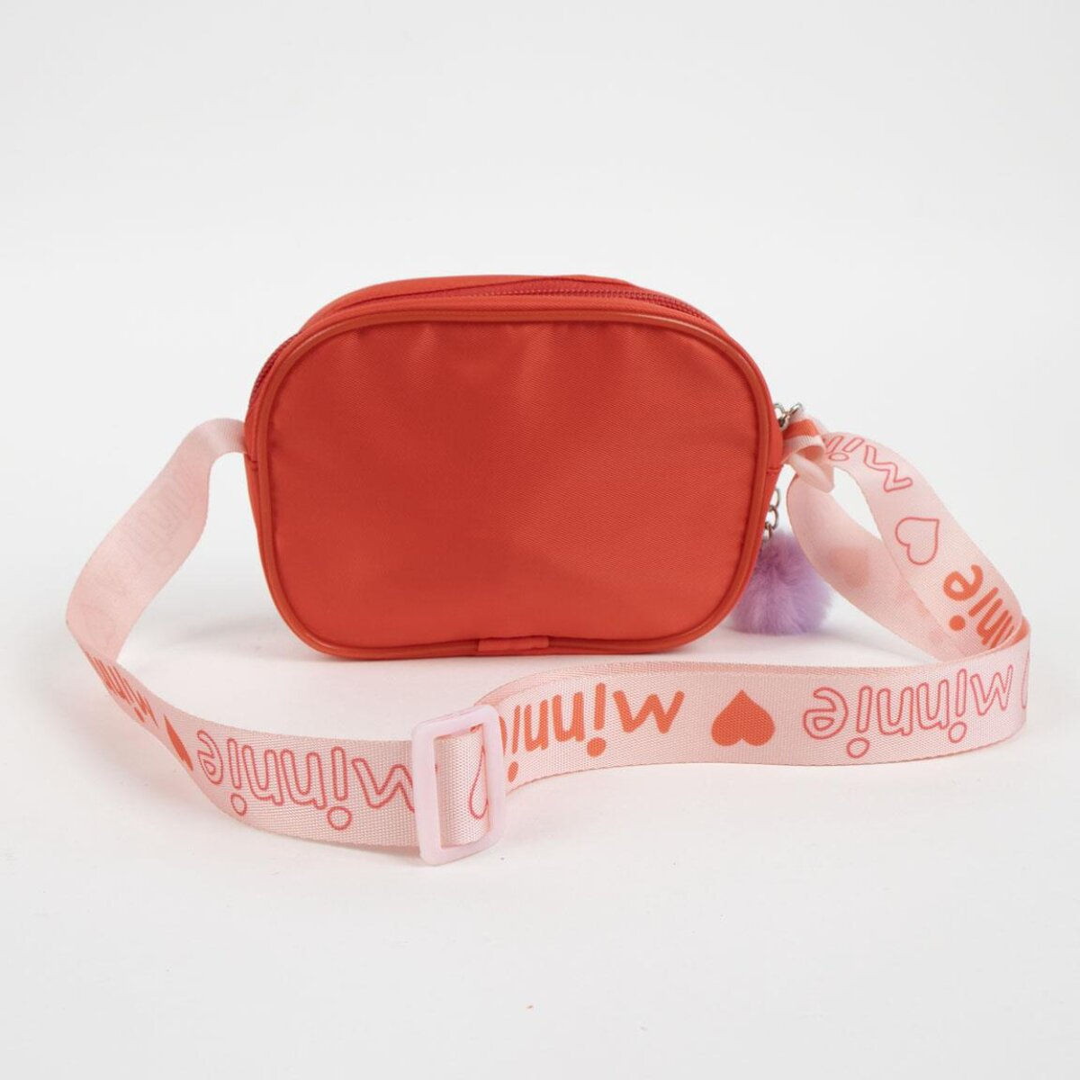 Un bolso rojo con una correa rosa. La correa dice 'Minnie' y tiene corazones. Un pompón morado cuelga del bolso.