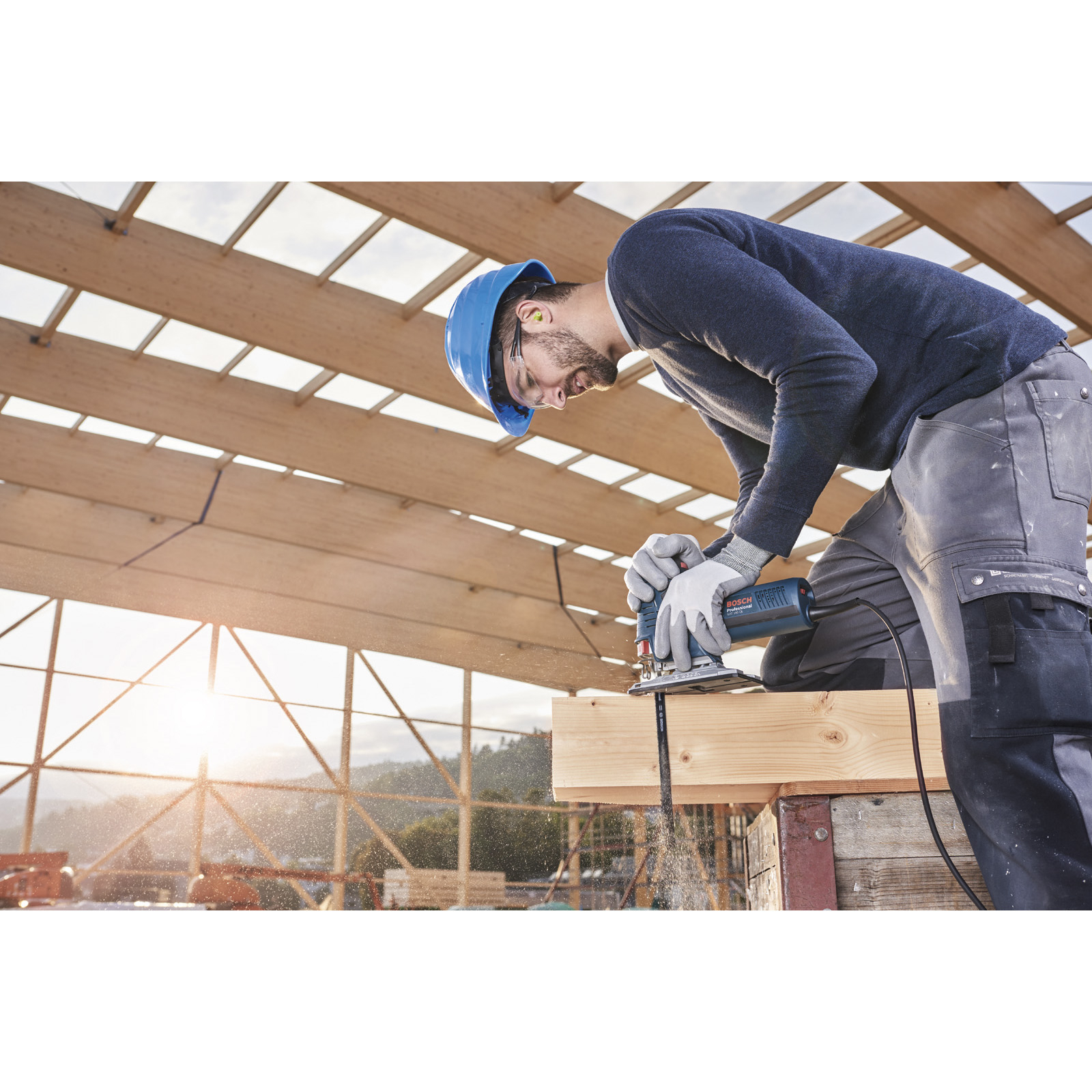 Stolarz używa wyrzynarki do drewna. Ma niebieski kask i okulary ochronne. Widoczne trociny i światło słoneczne.