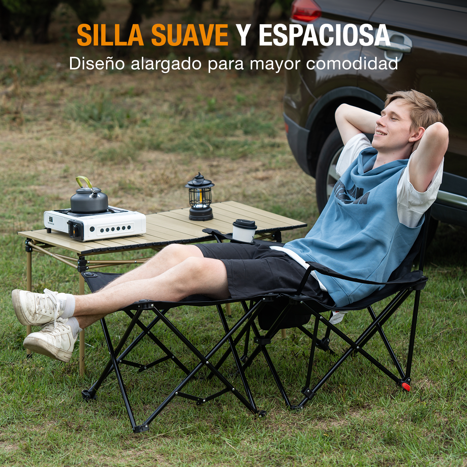 Hombre relajándose en silla negra. Mesa, estufa, linterna y coche al fondo. Hierba verde.