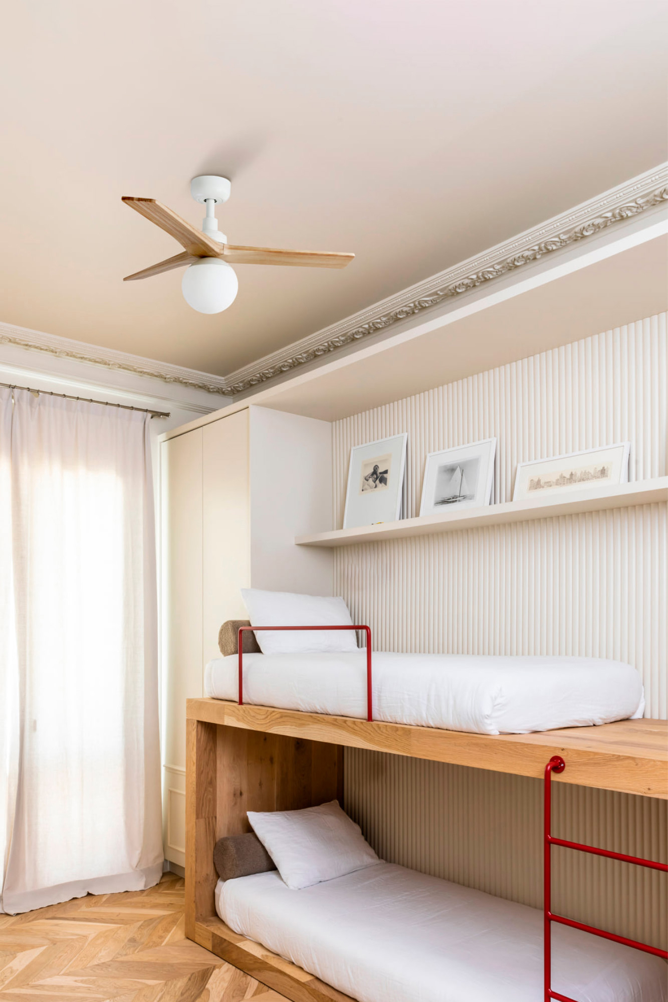 Ein Etagenbett mit weißer Bettwäsche und roten Akzenten. Darüber hängt ein Deckenventilator. Der Raum hat einen Holzboden und weiße Wände.