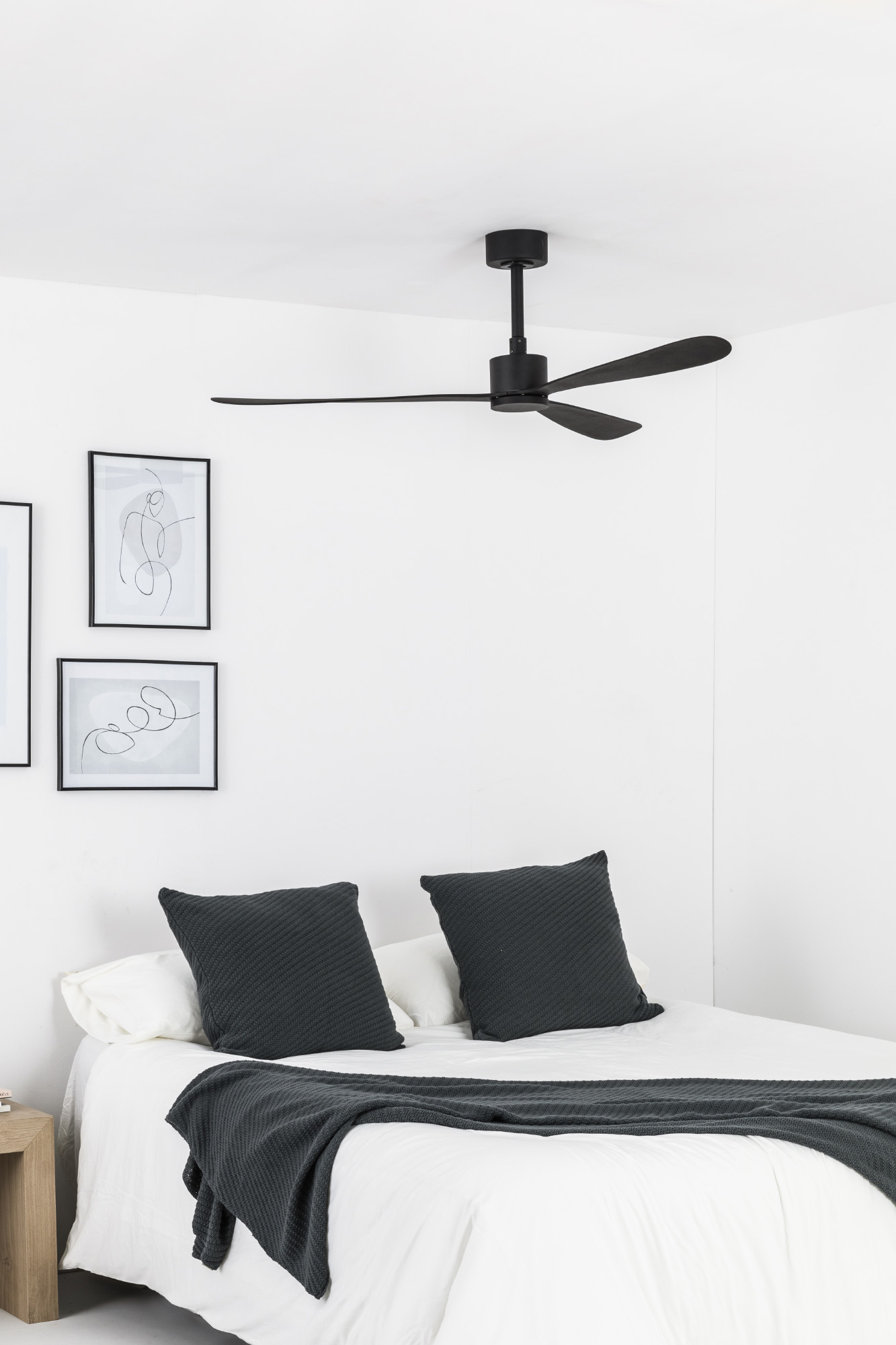 Una camera da letto con letto, cuscini, coperta e ventilatore a soffitto. Opere d'arte appese al muro bianco.