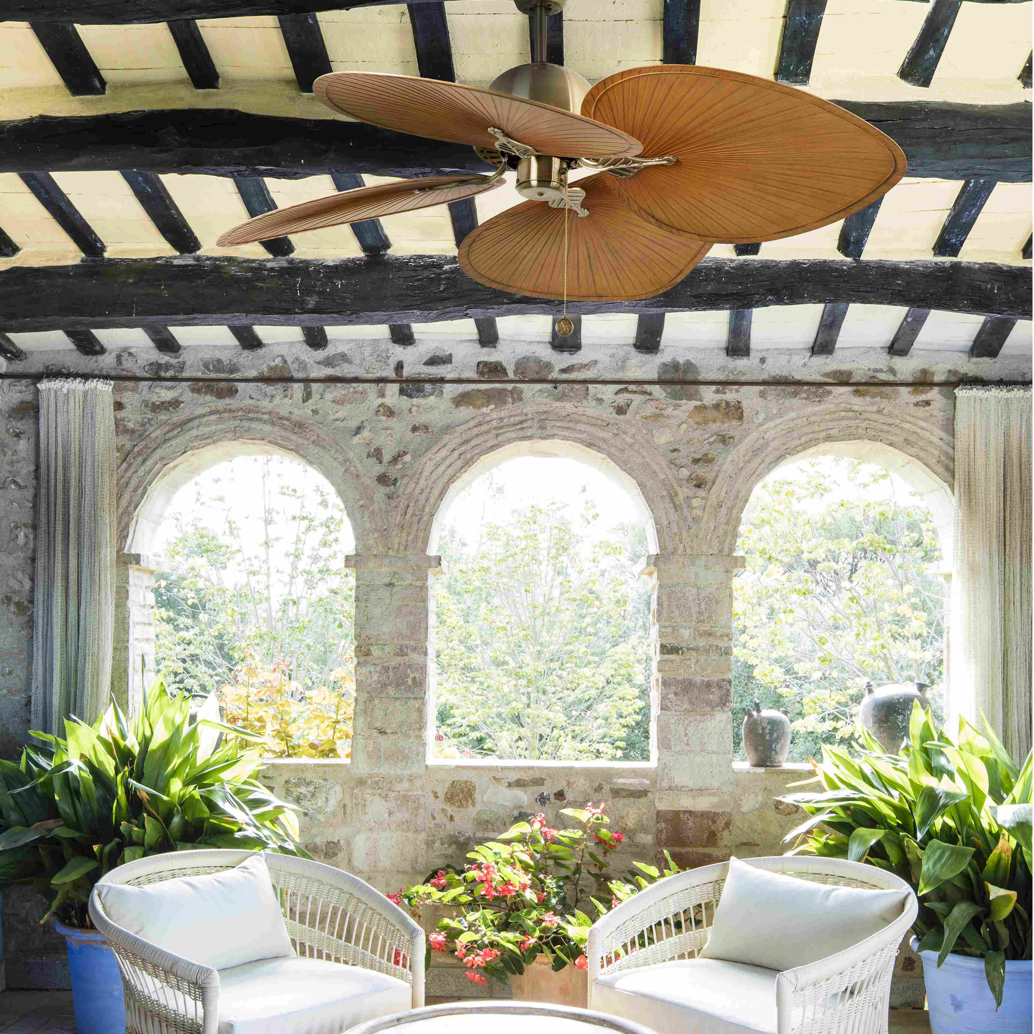 Una veranda con ventilatore a soffitto, muri in pietra, finestre ad arco, sedie bianche e piante. Vista giardino.