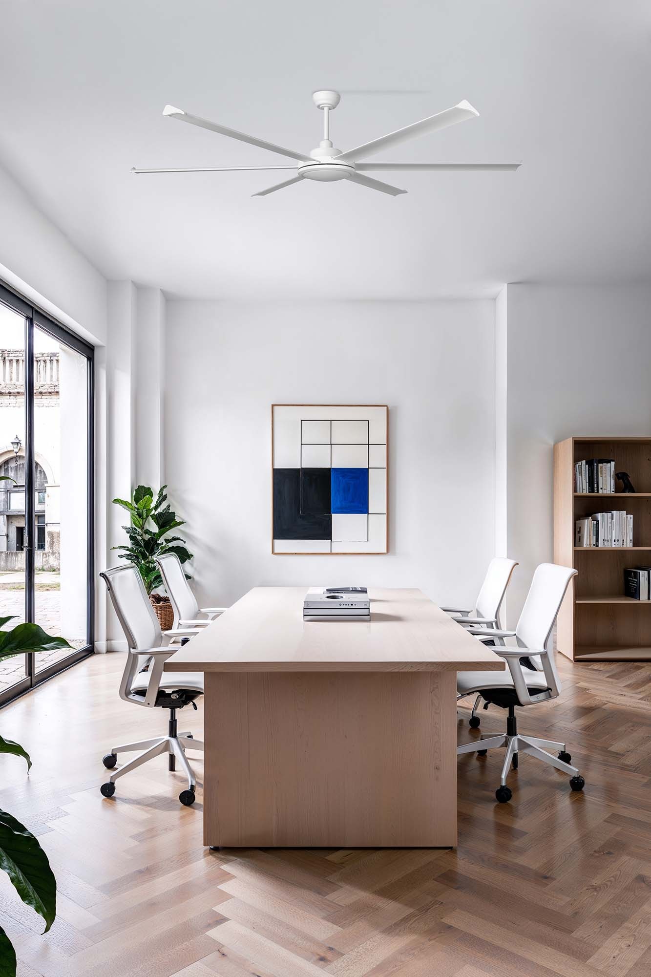 Ufficio moderno con un grande tavolo, sedie, opere d'arte e un ventilatore a soffitto. Luce naturale inonda la stanza.
