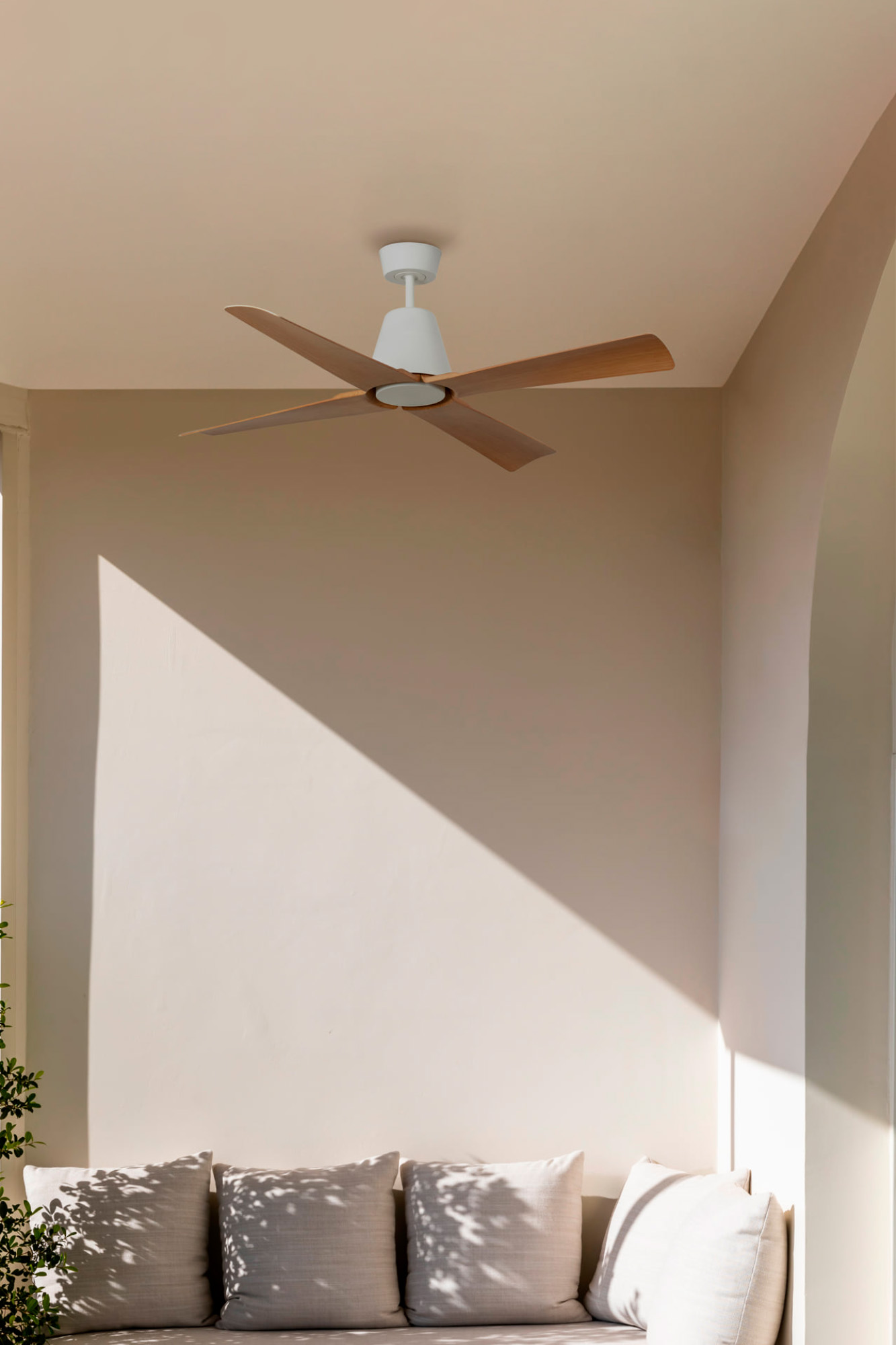 Ein Deckenventilator mit Holzflügeln hängt über einer beigefarbenen gepolsterten Bank. Sonnenlicht und Schatten erzeugen ein Muster an der Wand.