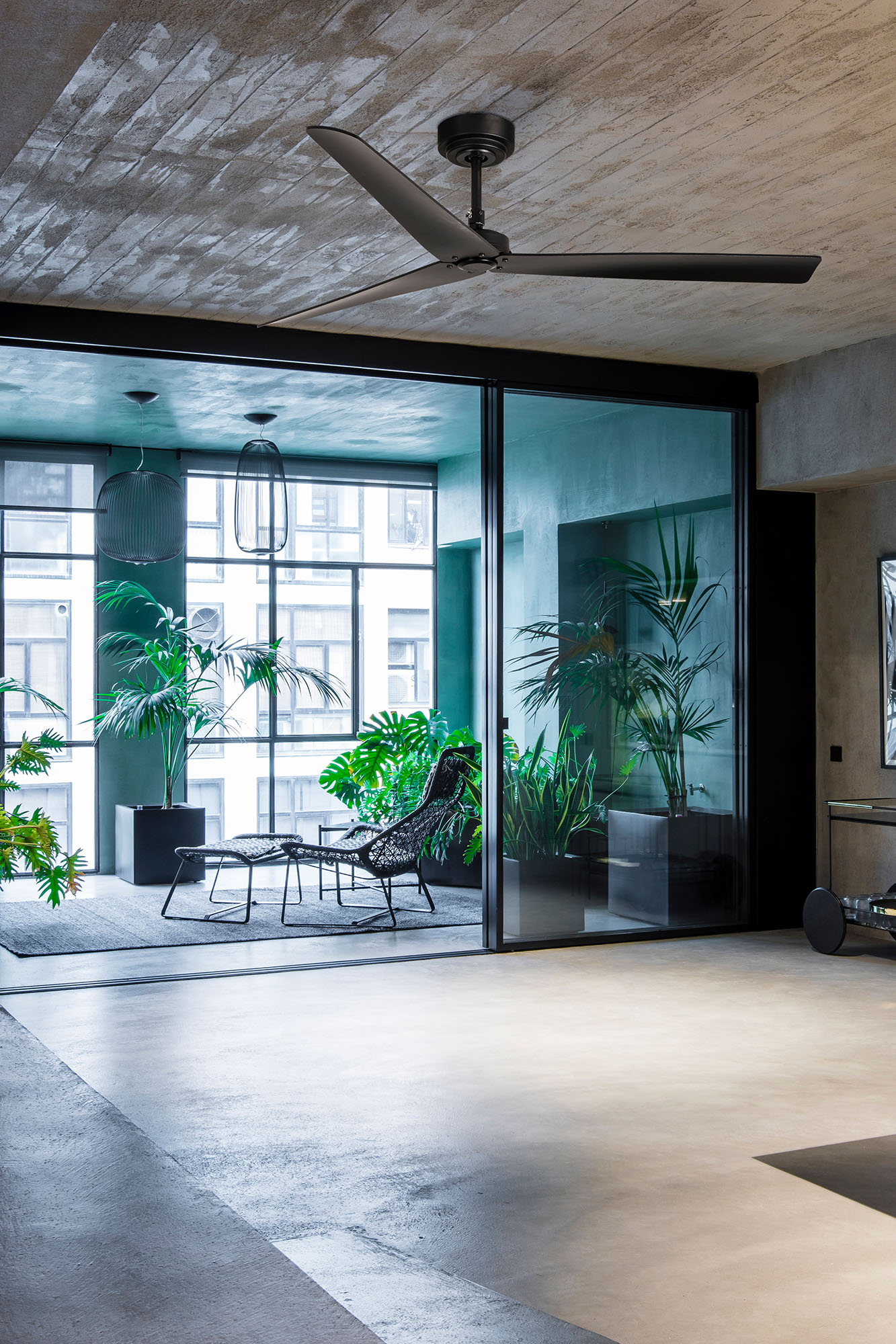 Modernes Interieur mit Pflanzen, schwarzem Deckenventilator, Glaswand und Sitzgelegenheiten im Freien. Betonboden und -decke. Helles, geräumiges Design.