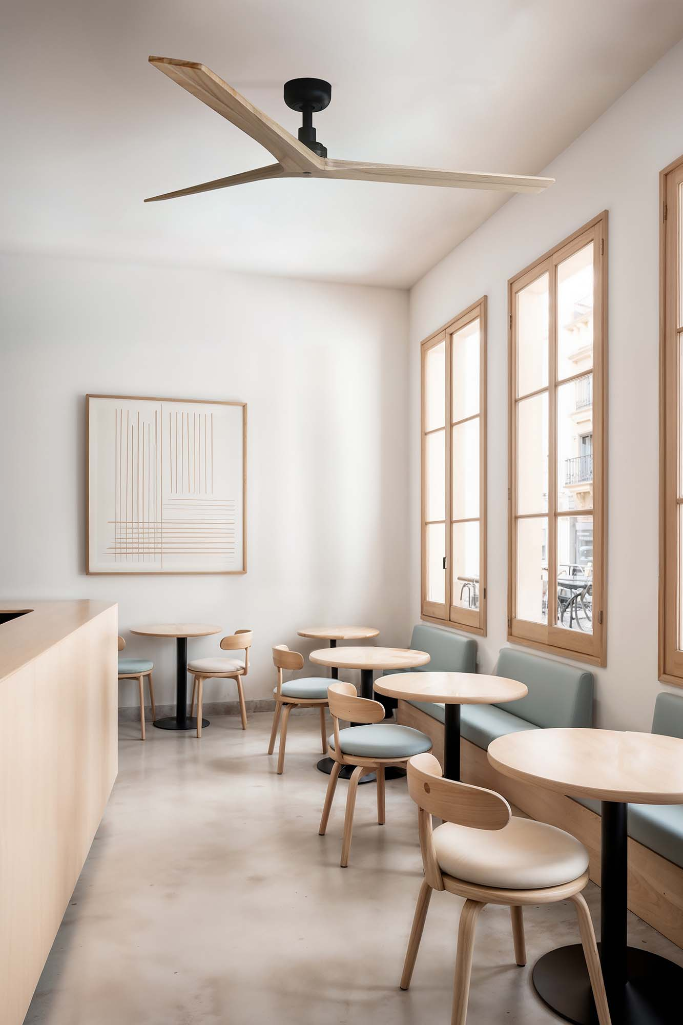 Interieur eines Cafés. Runde Tische, Holzstühle, eine Bank und Fenster. Ein großer Deckenventilator ist ebenfalls vorhanden.