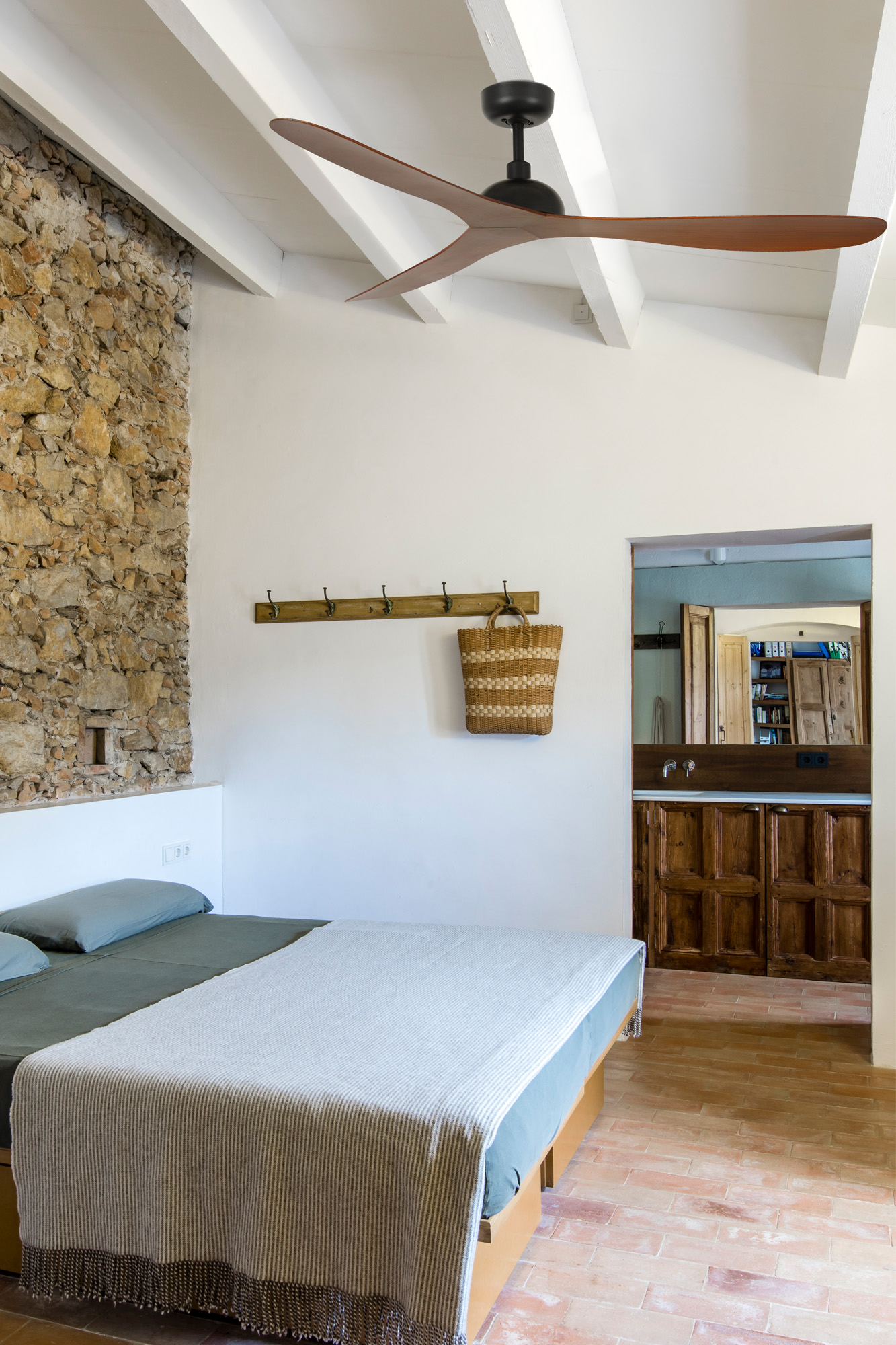 Camera da letto con muro in pietra, soffitto bianco con ventola, letto con coperta e vista su un bagno con elementi in legno.