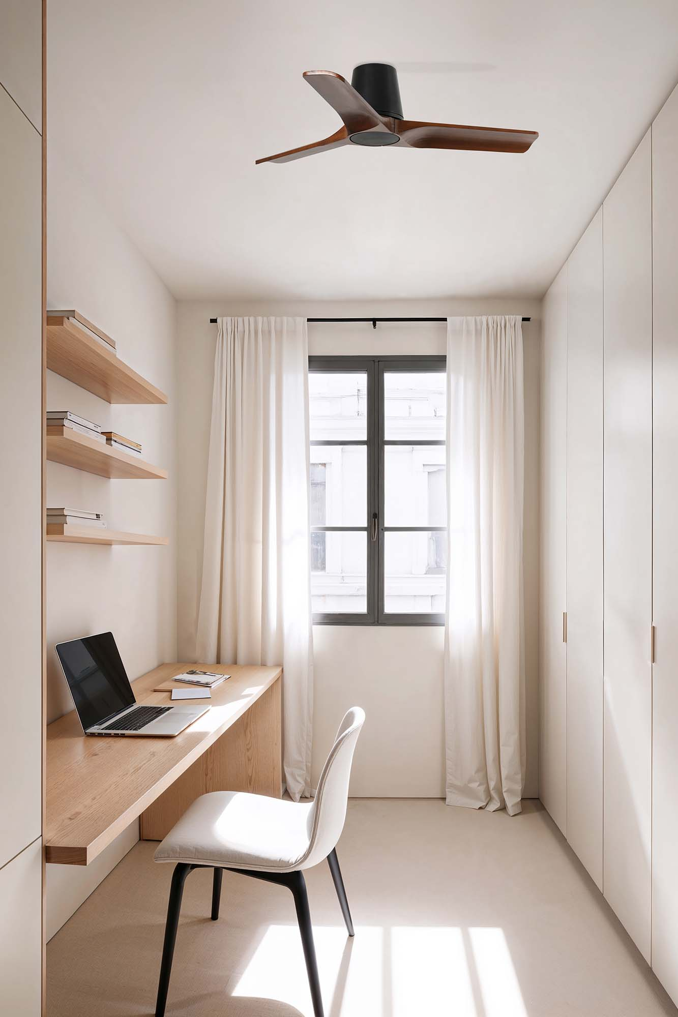 Ein modernes Heimbüro mit Schreibtisch, Stuhl und Deckenventilator. Ein Fenster mit weißen Vorhängen bietet Tageslicht.