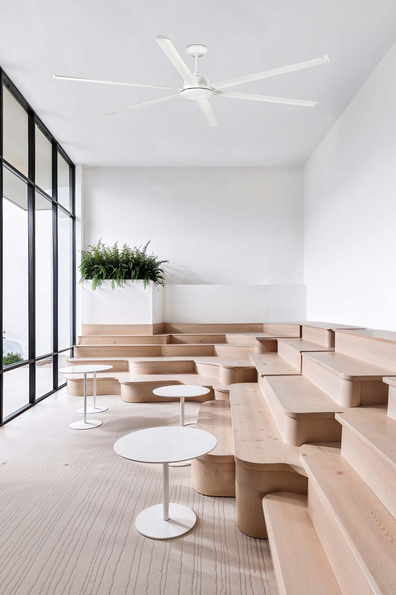 Un interno moderno con sedute in legno a gradoni, tavoli bianchi e una grande finestra con vista sull'esterno. Un ventilatore a soffitto è appeso sopra.