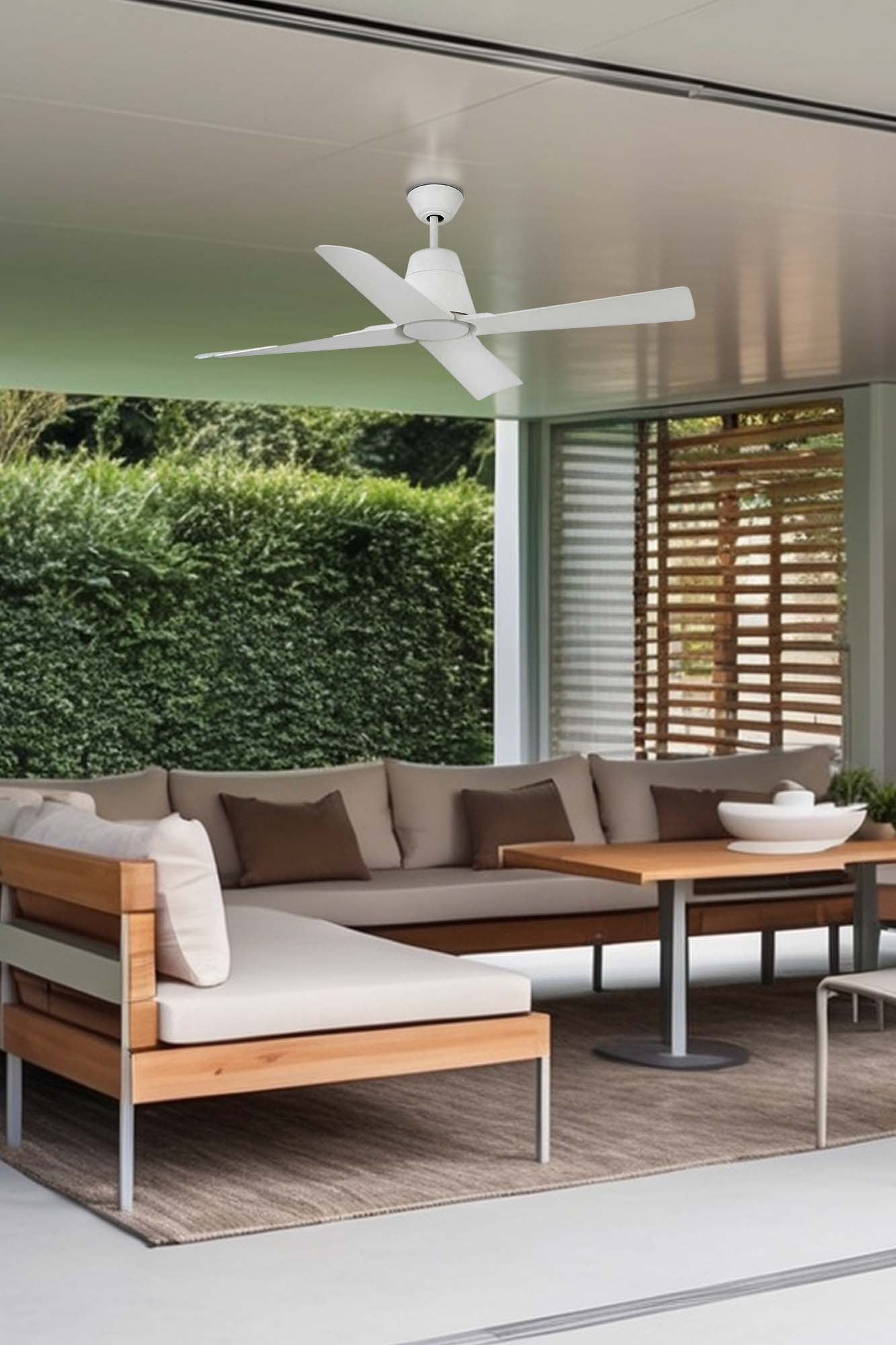 Un ventilatore a soffitto bianco è appeso sopra una moderna area salotto all'aperto. Una siepe verde è sullo sfondo. Un tavolo e un divano sono in primo piano.