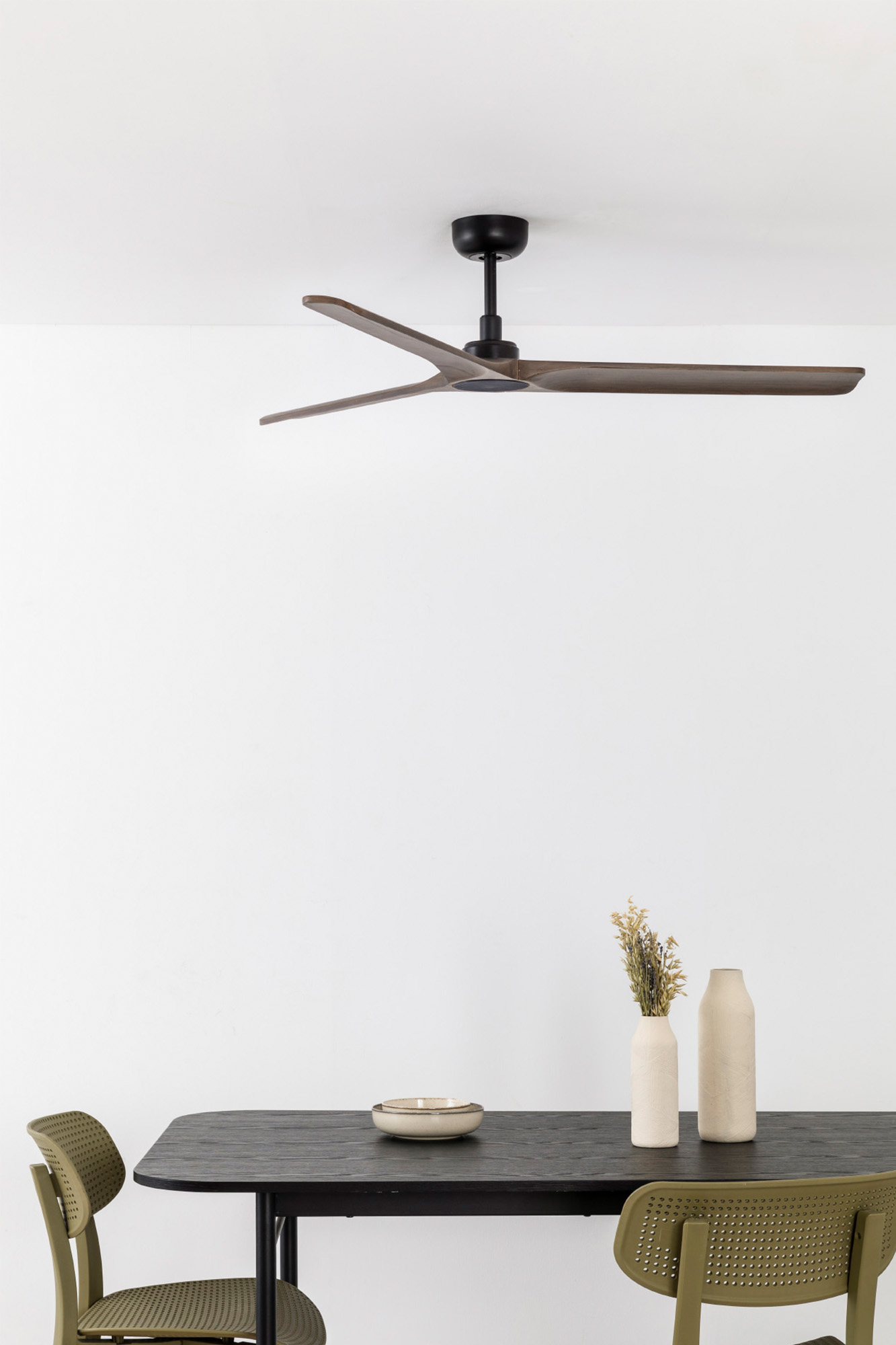 Ein Deckenventilator mit Holzflügeln hängt über einem dunklen Tisch mit Vasen und einer Schüssel. Zwei grüne Stühle stehen am Tisch.