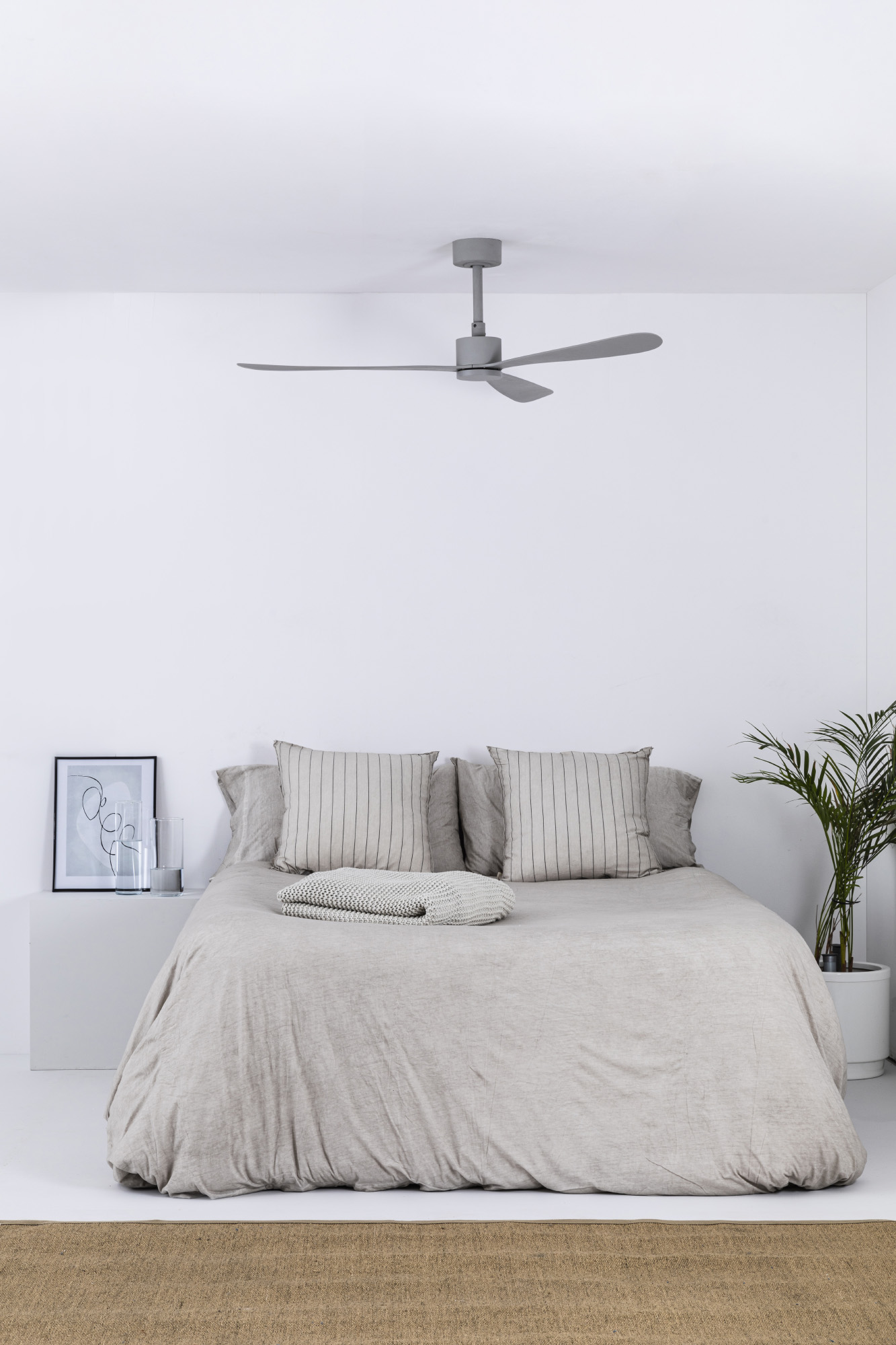 Schlafzimmer mit Bett, Kissen und Bettdecke in neutralen Tönen. Ein Deckenventilator und ein kleiner Tisch mit Bild und Glas.