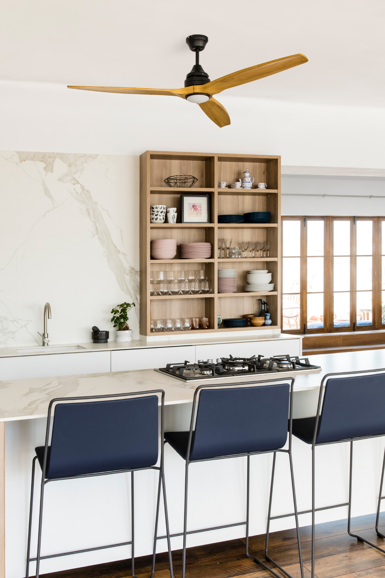 Cucina moderna con ventilatore a soffitto in legno, mensole con piatti, piano di lavoro in marmo e sgabelli blu navy.