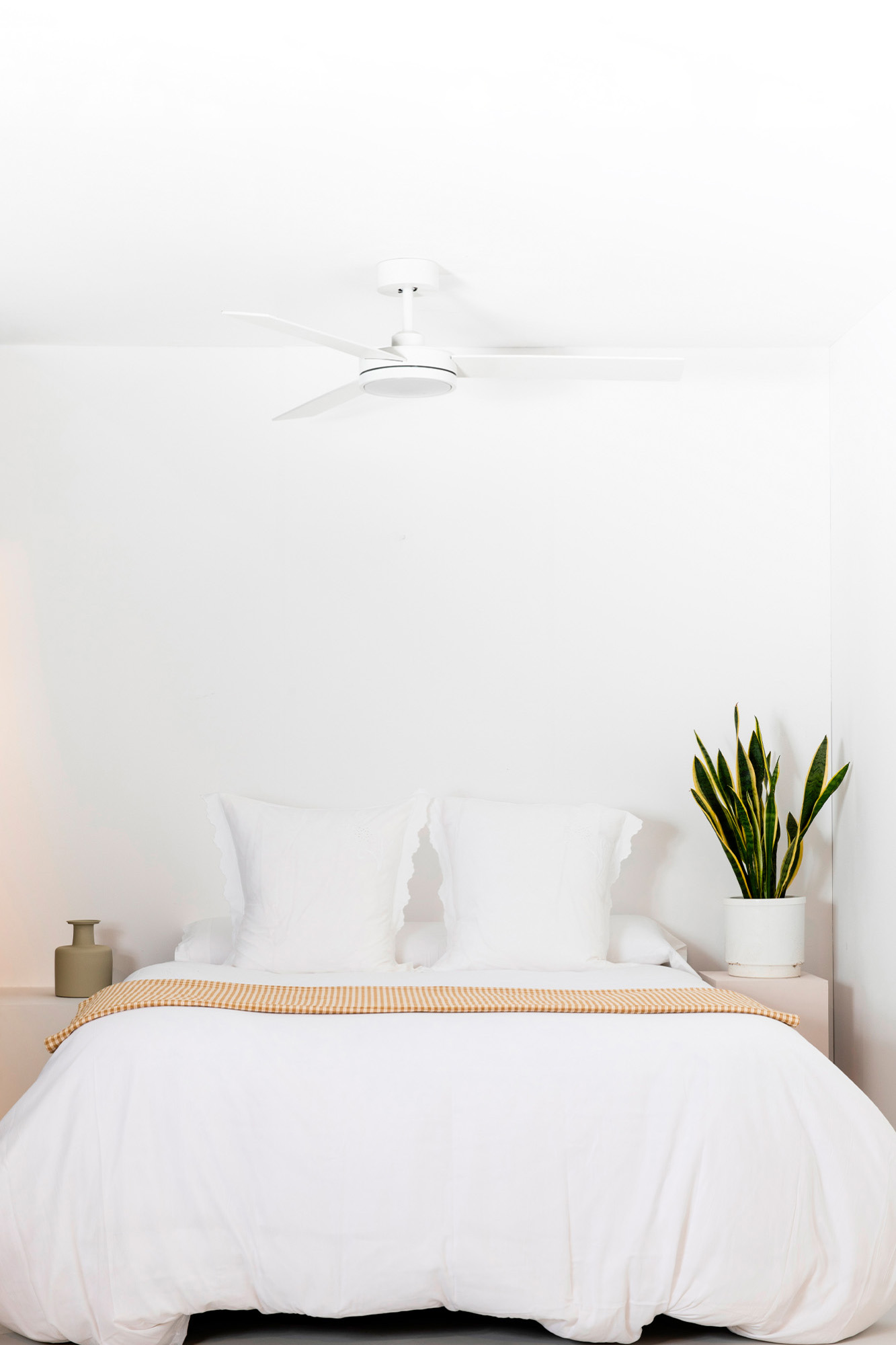 Una camera da letto bianca con letto, cuscini e pianta. Un ventilatore a soffitto è appeso sopra. Il letto ha una coperta gialla e bianca.