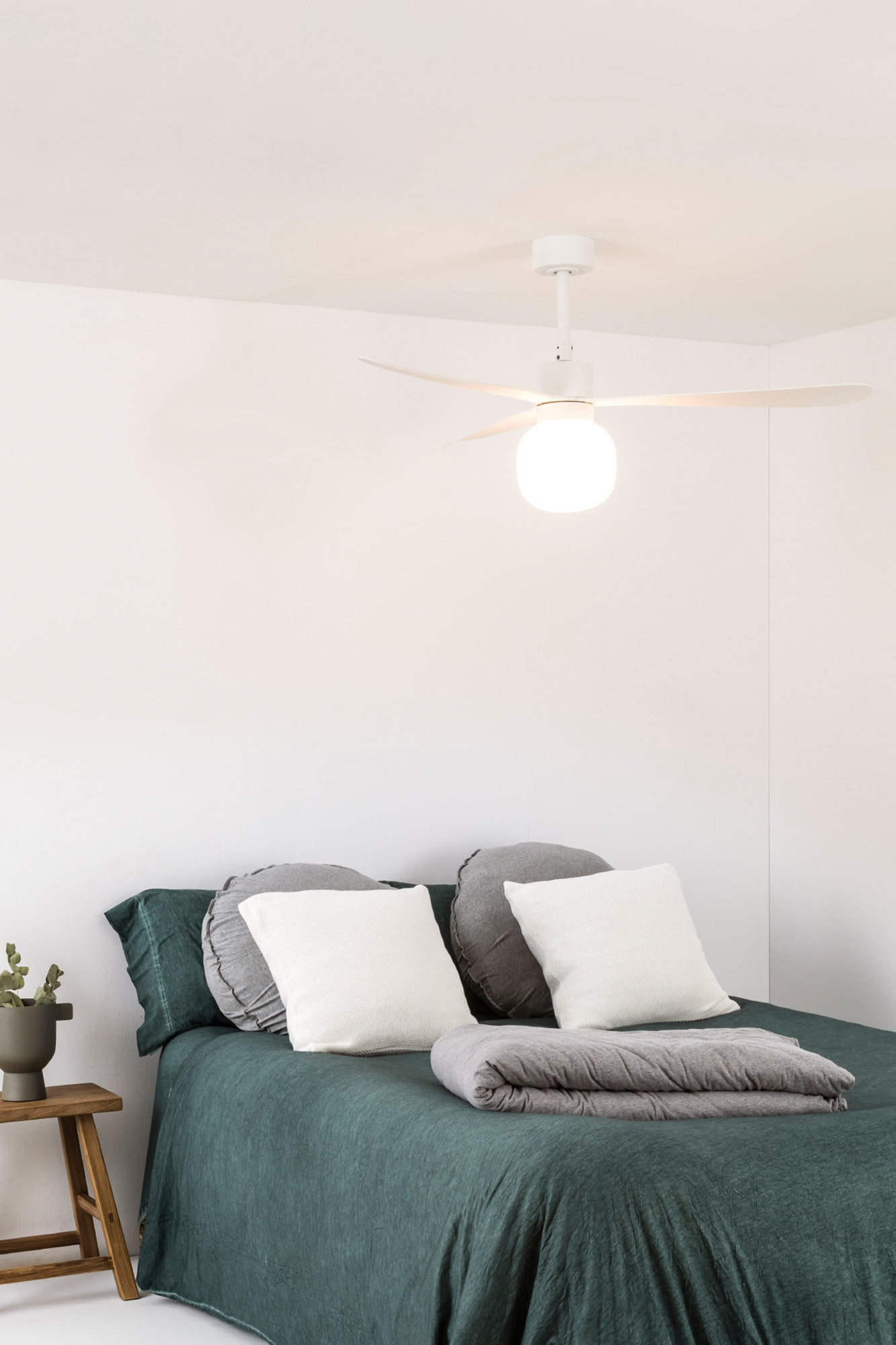 Ein Bett mit grüner Bettwäsche, weißen Kissen und einer grauen Decke. Ein Deckenventilator hängt oben. Links steht ein kleiner Holzschemel.