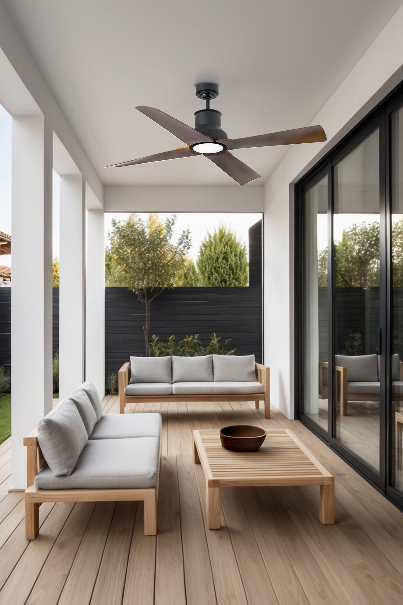 Patio esterno con mobili in legno, ventilatore a soffitto e porte scorrevoli in vetro. Lo sfondo mostra alberi e una recinzione nera.