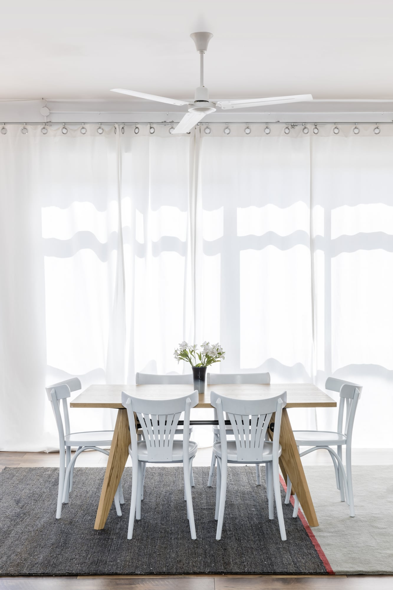 Una sala da pranzo con tavolo in legno, sedie bianche, tende bianche, ventilatore a soffitto e vaso di fiori.