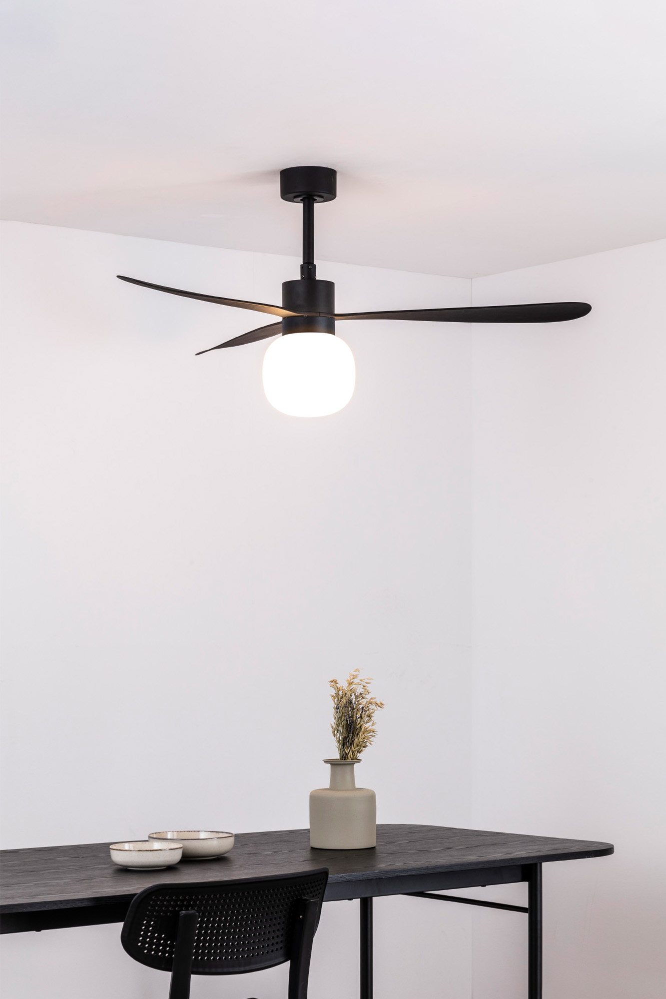 Ein schwarzer Deckenventilator mit Licht, Tisch mit Vase und Blumen und ein schwarzer Stuhl. Weiße Wände.