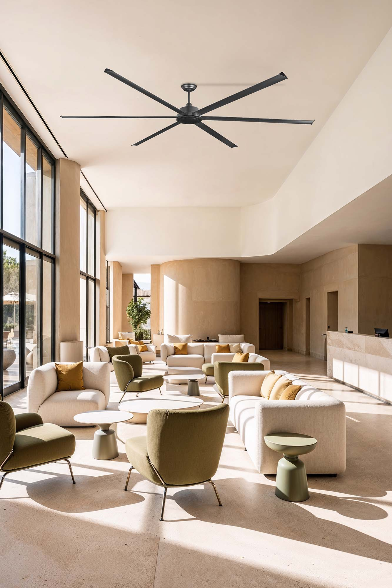 Ampia hall con grandi finestre, divani, poltrone e un ventilatore a soffitto. I colori sono beige, verde oliva e bianco.