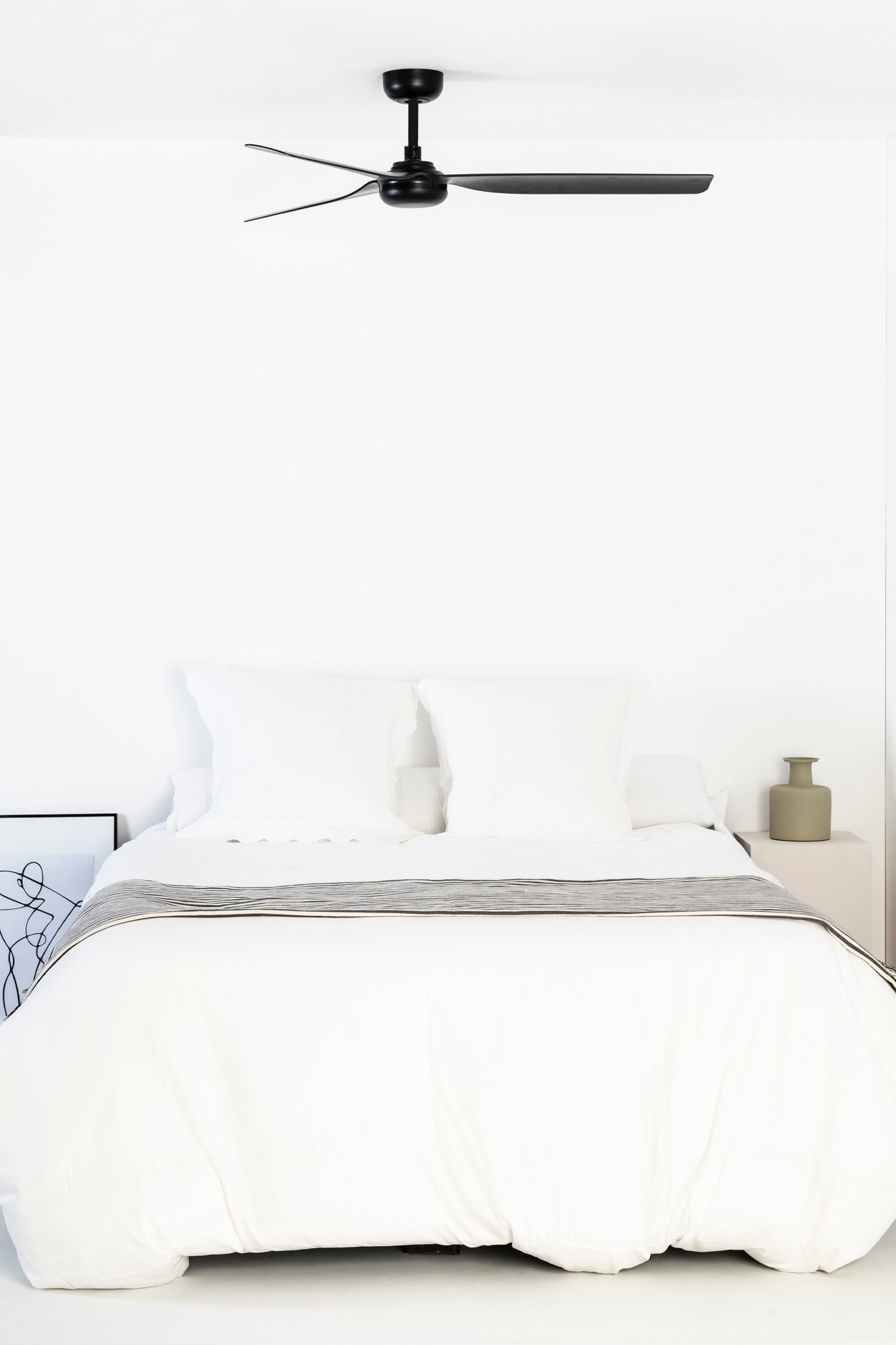 Una camera da letto minimalista con letto bianco, cuscini e ventilatore a soffitto nero. Un vaso è su un comodino.